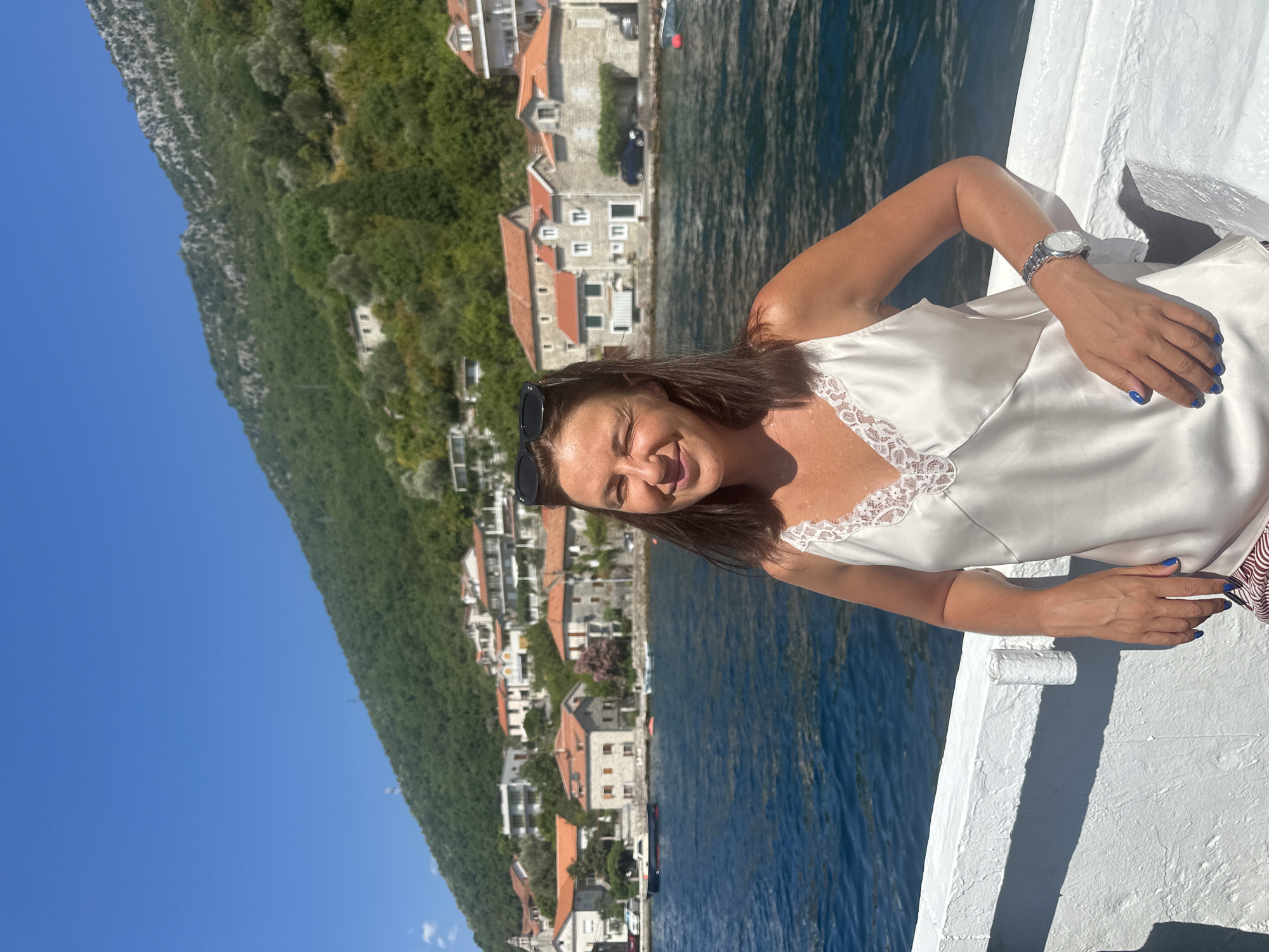 A woman with dark brown hair and sunglasses perched on her head smiles at the camera while leaning on a white railing. She is wearing a white satin camisole with lace trim and striped pants. Her left hand rests on the railing, displaying blue nail polish and a wristwatch. Behind her, the deep blue water of the Bay of Kotor extends towards a village in Lepetane, Montenegro. The village features traditional stone houses with terracotta roofs nestled on a steep, green hillside. The sky is clear and blue. The woman appears to be on a boat, as evidenced by the white railing.