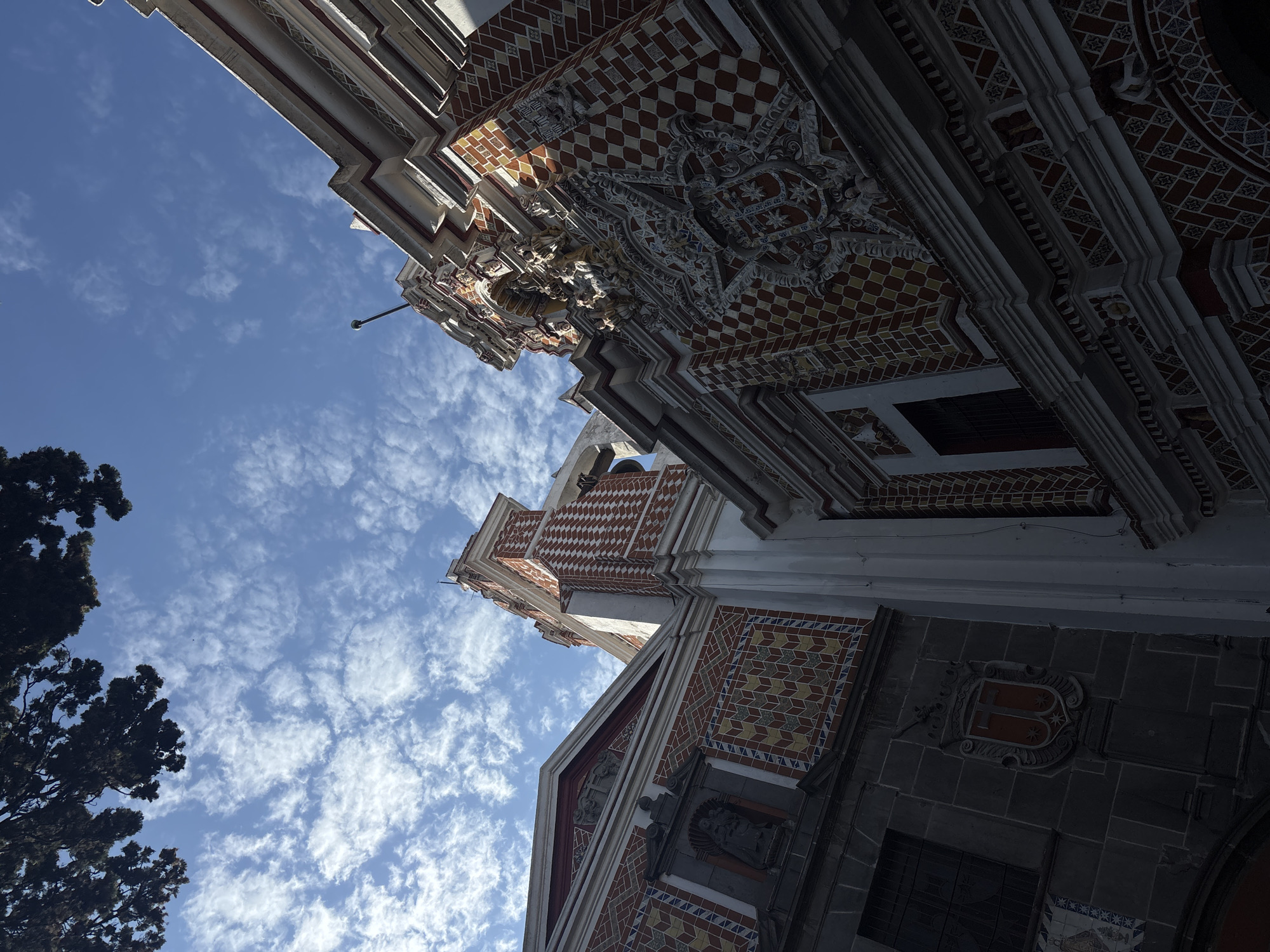 The image captures a low-angle view of the facade of a church, likely the Templo de San Francisco Acatepec in Puebla, Mexico, known for its intricate Talavera tilework. The facade features a mix of geometric tile patterns in colors like red, yellow, and white, along with ornamental carvings and sculptures. The architecture is predominantly Baroque. The image also includes a section of sky with light cirrocumulus clouds and the top of a tree in the left corner. The light suggests it is daytime. No people are visible.
