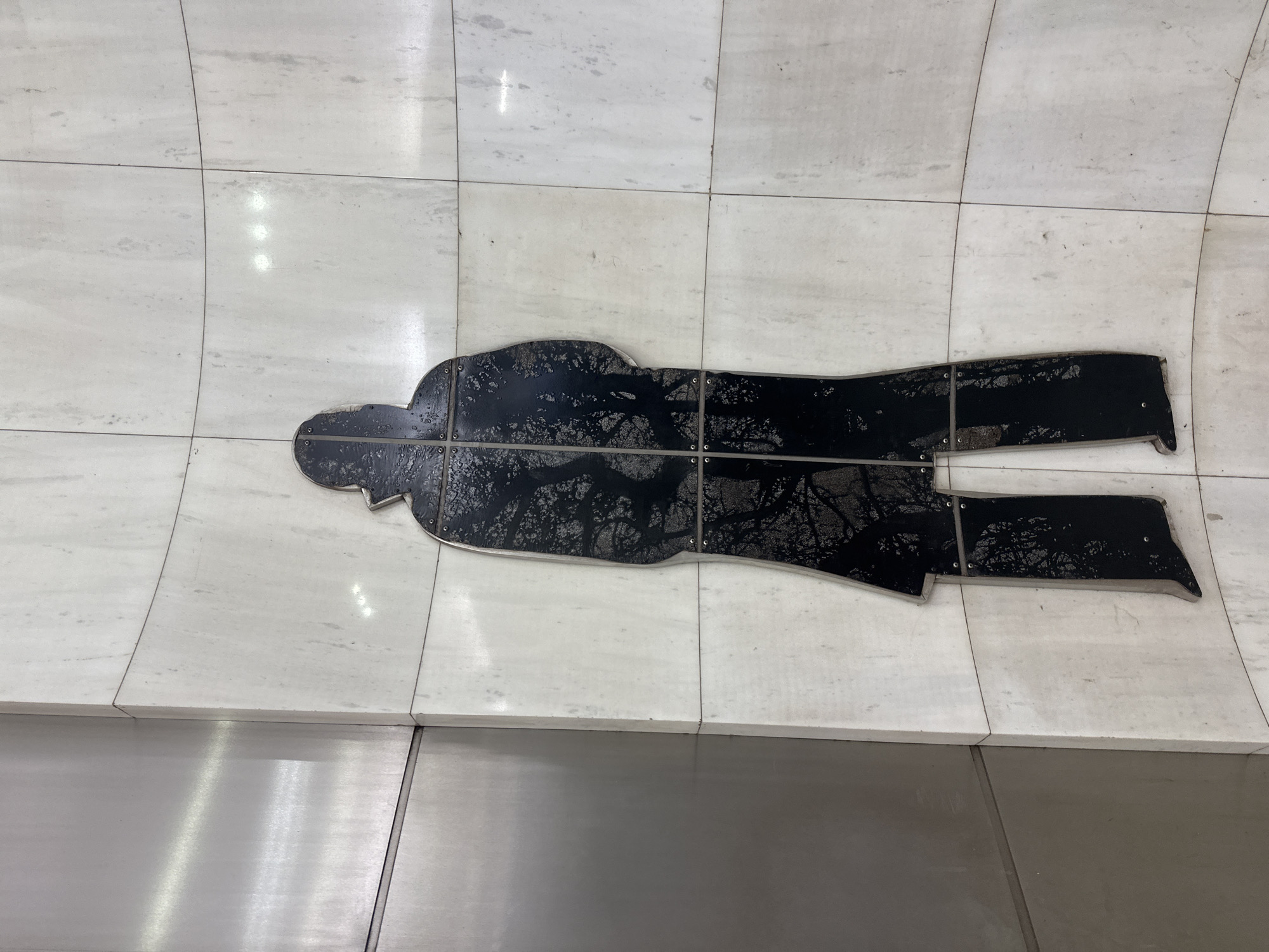 This is a full shot of a silhouetted figure on a marble wall, likely in Moscow, Russia. The silhouette is of a person wearing a long coat and a hat, facing away from the viewer. The silhouette is filled with an image of tree branches, creating a stark contrast against the white marble wall. The wall is made of large, square tiles with thin grout lines. To the left of the silhouette, there is a brushed metal panel. The lighting suggests this is an indoor environment, possibly a public space like a subway station or a museum. The silhouette is attached to the wall with small, visible screws.