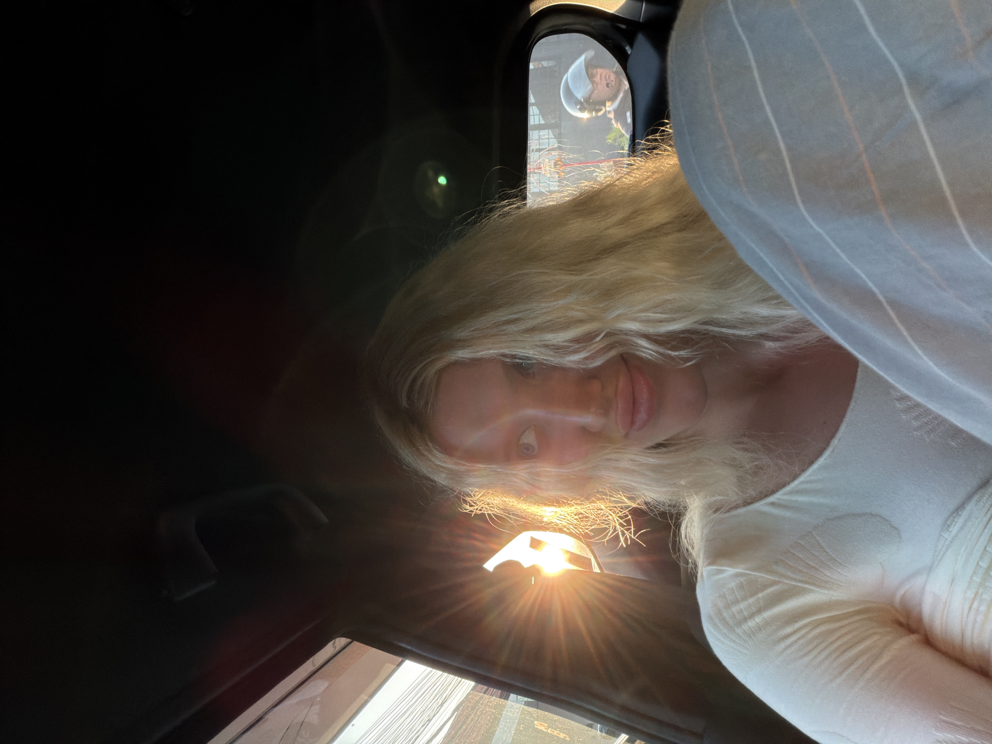 The media file shows a close-up, slightly tilted selfie of a person with long, blonde hair inside a vehicle. The person is looking towards the camera with a gentle smile. Sunlight is directly hitting the lens, creating a lens flare and a bright glow. In the rearview mirror, another person is visible, wearing a light-colored hat and a dark jacket. The setting appears to be during the daytime, likely late afternoon given the angle of the sun. The interior of the car is visible, including a portion of the passenger seat or clothing with a striped pattern. The background outside the car is blurred but suggests an urban or suburban environment, possibly with buildings or vegetation. There is no visible text in the image. The overall mood seems relaxed and casual.