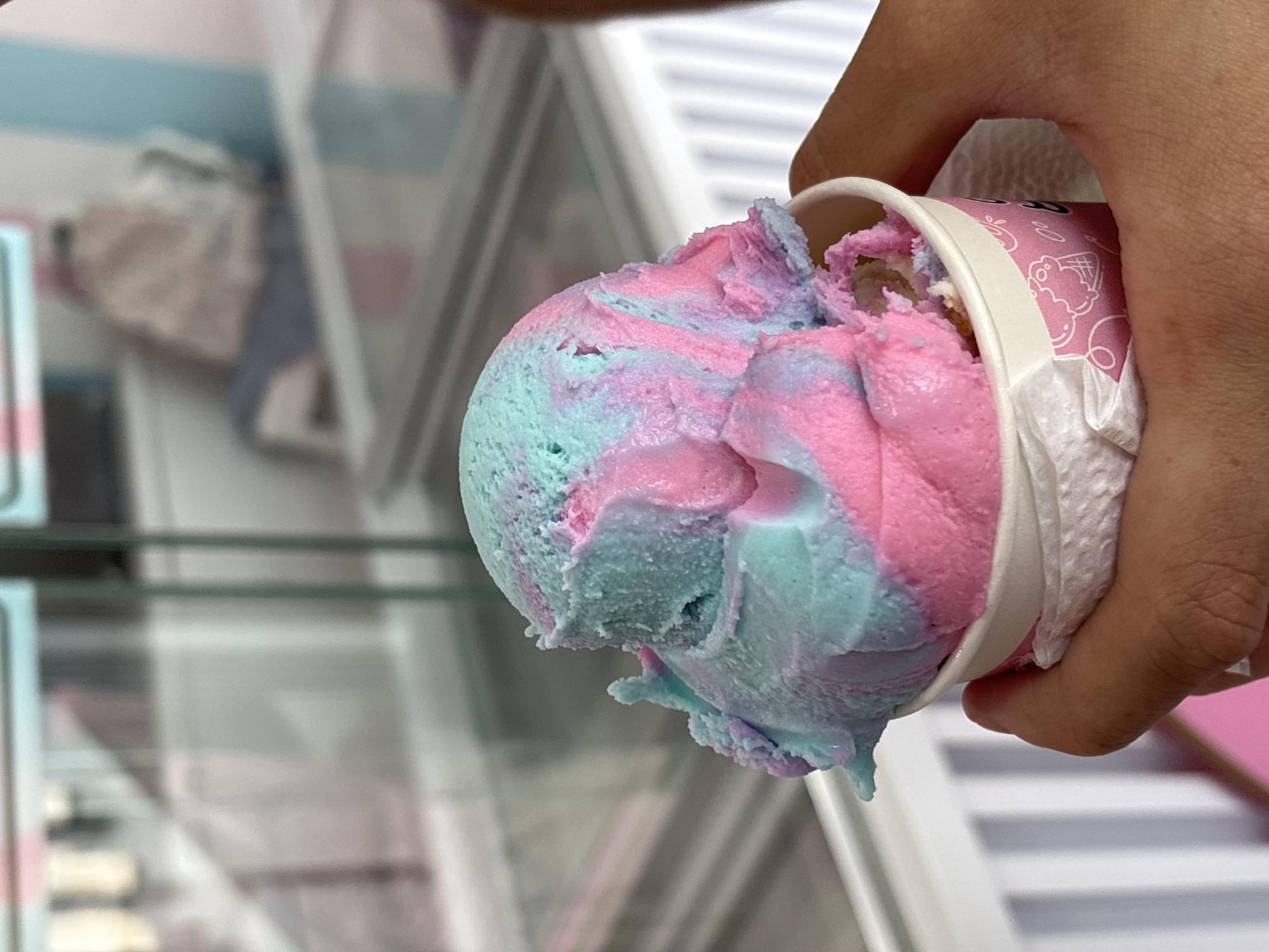 A close-up shot shows a hand holding a paper cup filled with multicolored ice cream. The ice cream is predominantly pink and blue, swirled together. The cup has a white rim and a pink exterior with cartoon illustrations. A white paper napkin is wrapped around the cup. The background is blurry but suggests a retail environment, possibly a display case or shelving unit. The image does not provide sufficient information to determine the specific location in Santa Clara Ocoyucan, Mexico.