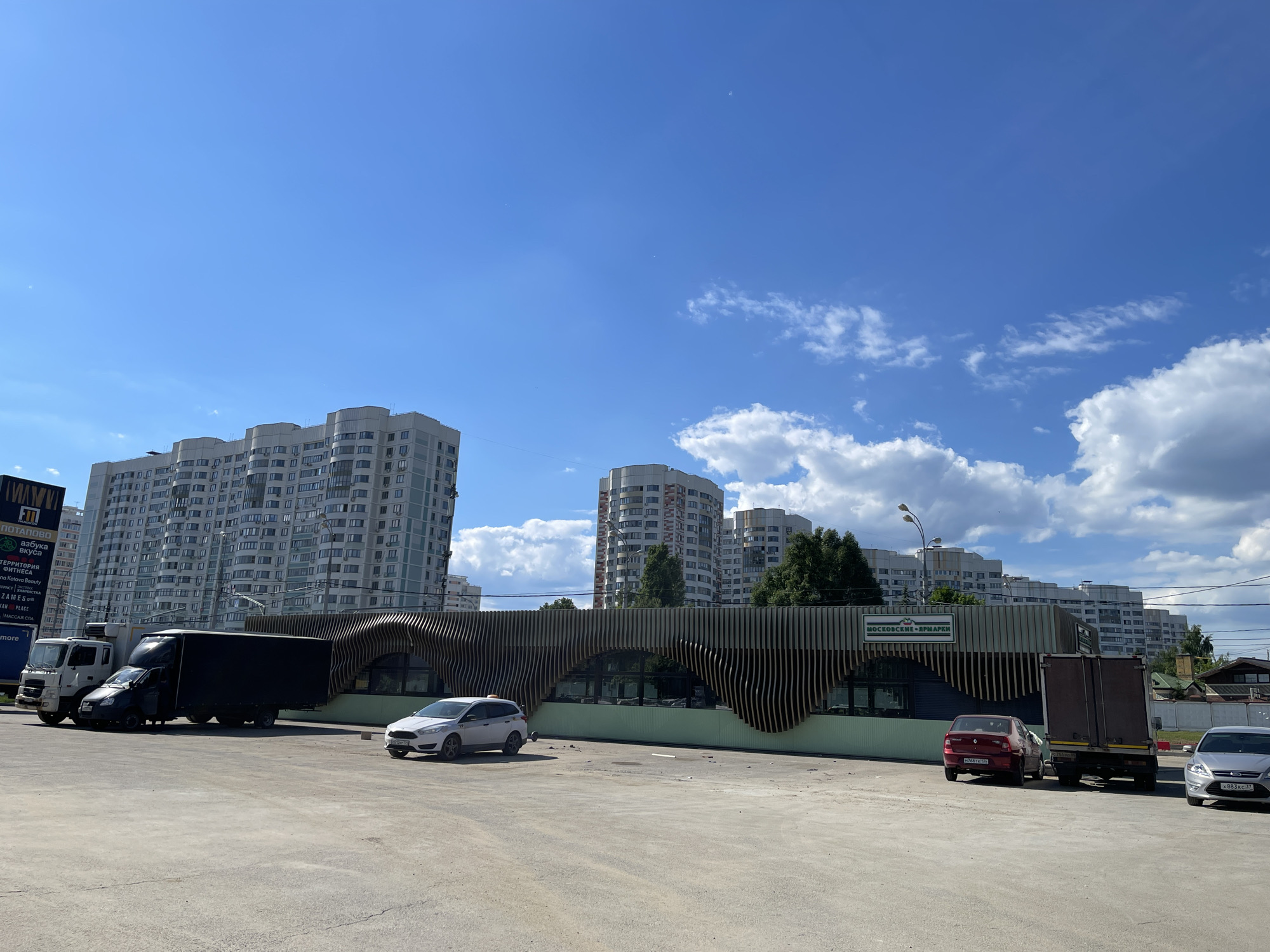 The image captures a vibrant urban scene in Moscow, Russia, under a clear blue sky with scattered white cumulus clouds, indicating a bright and sunny day. In the background, several modern, light-colored apartment buildings with numerous windows and balconies rise, showcasing contemporary residential architecture.
In the midground, a distinctive low-rise commercial building features a unique wave-like facade made of vertical wooden slats, partially painted green at its base. A prominent sign on this building reads "МОСКОВСКИЕ ЯРМАРКИ" (Moscow Fairs).
The foreground is a large, paved parking area where several vehicles are parked. These include a white and black truck, a dark-colored hatchback car, a white Ford Focus hatchback, a red Renault Logan sedan, and another dark truck with a box trailer, along with parts of another silver car. On the far left, a tall vertical sign board is visible, listing various establishments: "ПОТАПОВО", "АЗБУКА ВКУСА" (Azbuka Vkusa), "ТЕРРИТОРИЯ ФИТНЕСА" (Territory of Fitness), "ЯНА KOTOVA BEAUTY", "ПТРАСТ ХИМЧИСТКА", "ZAMES", "KAH PLACE", and "МАССАЖ СПА".
The scene depicts a typical commercial and residential area, with no people visible. The activity is limited to parked vehicles, suggesting an ordinary daytime scene in the city.
