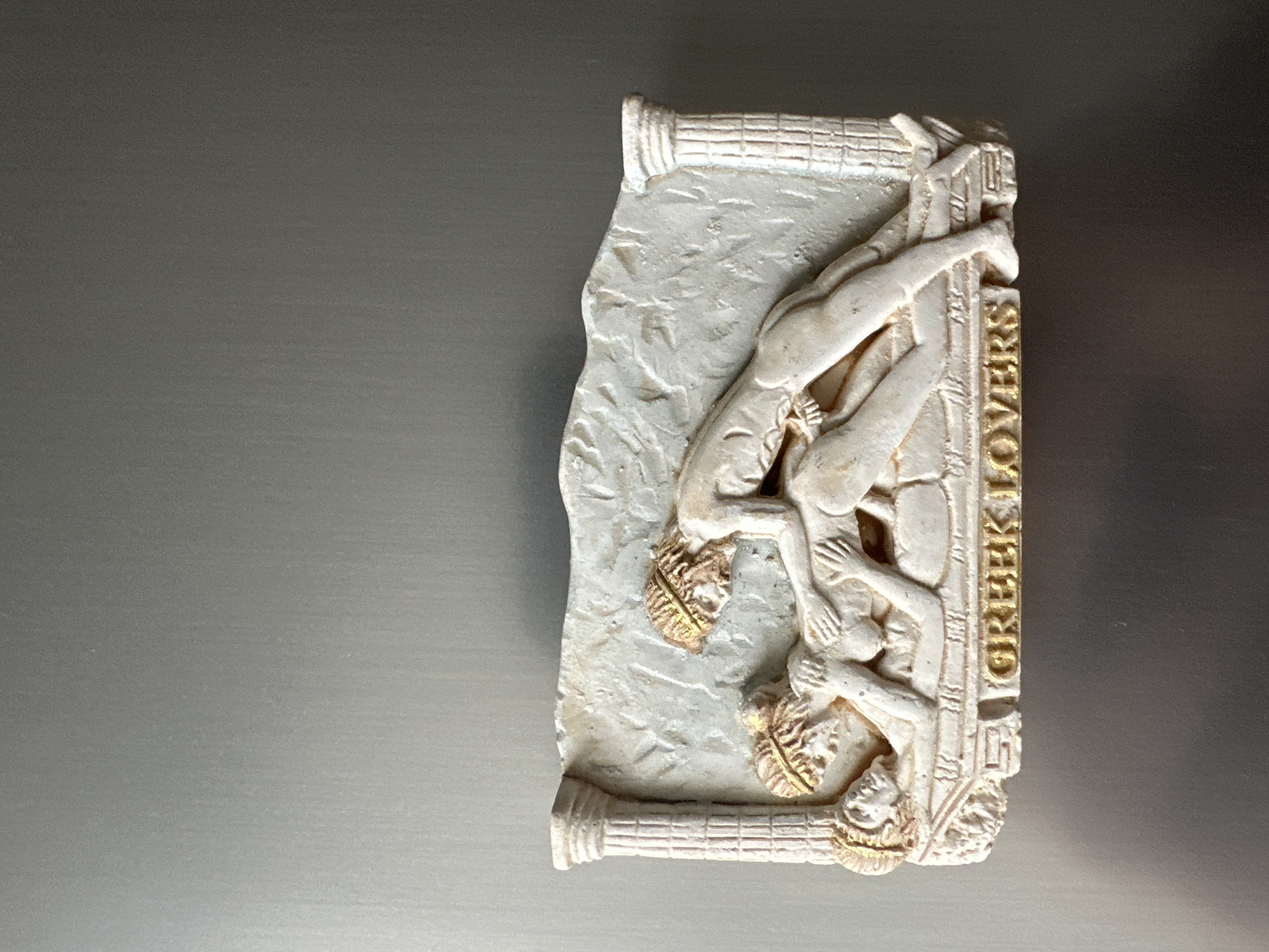 A white rectangular relief magnet featuring two figures intertwined in an embrace is affixed to a brushed metal surface, possibly a refrigerator. The figures, rendered in a classical style with golden headbands, are depicted on a textured background with two fluted columns framing the scene. Below the figures, the words "GREEK LOVERS" are inscribed in raised gold lettering. The overall color palette consists of beige, white, and gold, set against the cool gray tones of the metallic background.