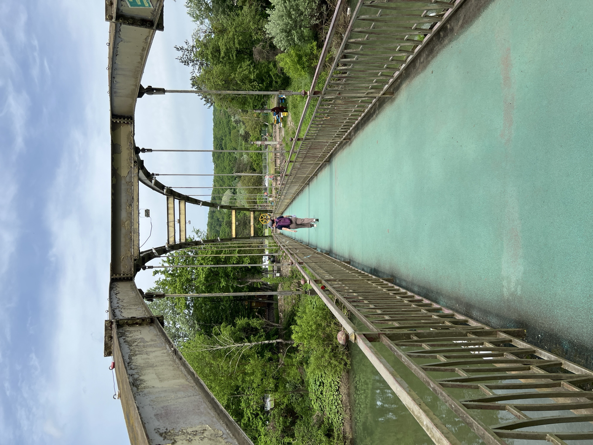 The image shows a suspension bridge in Goryachiy Klyuch, Russia. The bridge has a green surface and metal railings. Two people are walking on the bridge, heading away from the camera. The bridge appears to be over a body of water. Lush green vegetation is visible on both sides of the bridge. The sky is overcast.