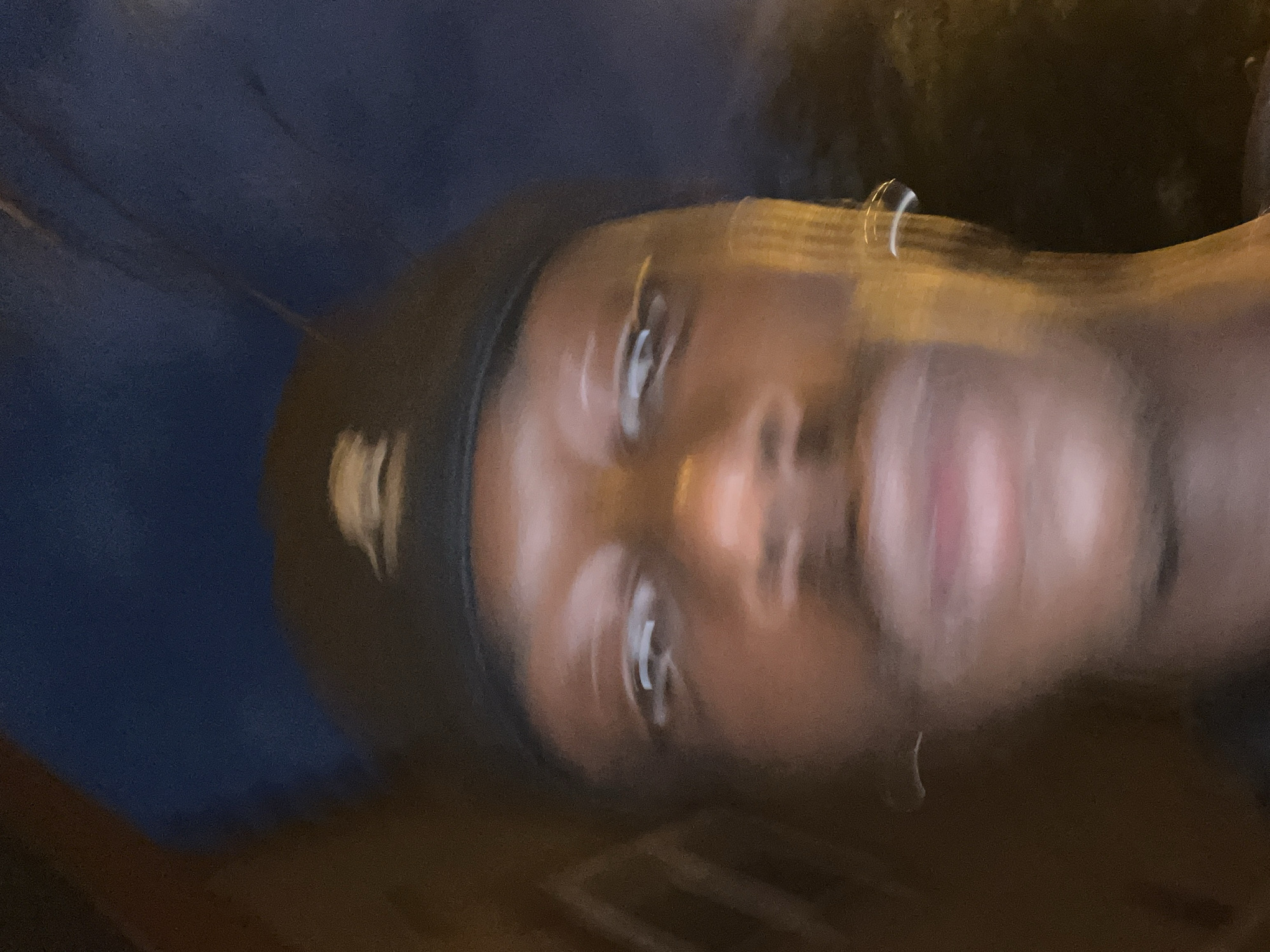 The image shows a person's face, possibly male, with dark skin and wearing a black head covering. The face is slightly blurred, and the background is out of focus, suggesting movement during the photo. There is no discernible information to indicate this image was taken in Barcelona, Spain. The surrounding environment is not clear enough to deduce a specific location.