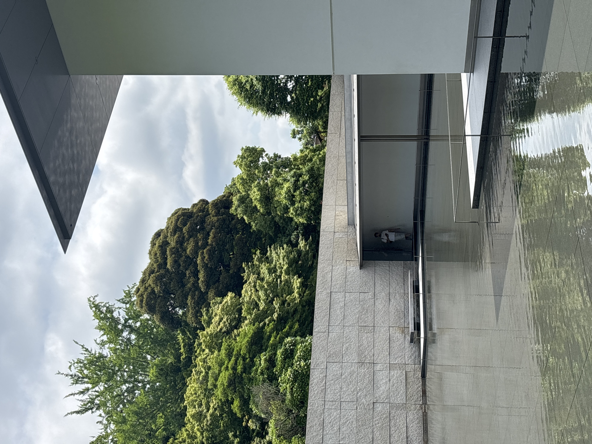 This image captures a view of the 21st Century Museum of Contemporary Art in Kanazawa, Japan. The scene features a modern architectural design, a reflecting pool, and dense natural foliage under a cloudy sky. A large, shallow reflecting pool occupies the lower half of the frame, its surface displaying ripples and reflections of the surrounding elements. To the far right, a tall, plain, light-colored building wall extends vertically, topped by a dark, flat, cantilevered roof overhang whose underside reveals a subtle grid pattern.
In the mid-left, a horizontal wall constructed from light grey, textured stone blocks provides a visual boundary. Set within this stone wall is a sheltered alcove or walkway, where a single individual dressed in light clothing stands, facing right. A minimalist bench or seating platform is positioned at the edge of the reflecting pool in front of the stone wall. Behind the architectural elements and dominating the upper left and central background, a dense expanse of lush green trees, displaying various shades from light to dark green, creates a vibrant natural backdrop. The sky above is overcast with soft, grey clouds.