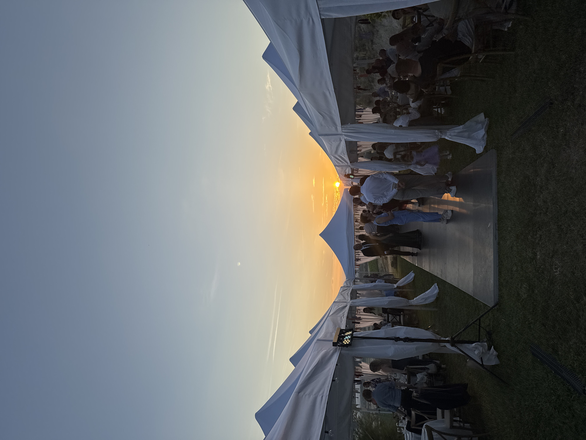 An outdoor event is depicted at sunset in Harsovo, Bulgaria. Two parallel rows of white, pointed-roof canopy tents are erected on a grassy field, framing a central, dark-colored elevated walkway or dance floor that extends towards the western horizon. The sky exhibits a gradient from light blue overhead to bright orange and yellow at the horizon, where the sun is partially visible. A faint crescent moon is discernible in the upper right quadrant of the sky, along with minimal wispy clouds and faint contrails.
Within the tents, guests are seated at tables adorned with white tablecloths and chairs, engaged in dining and conversation. On the central walkway, multiple individuals are standing and mingling; this group includes adults in semi-formal attire and at least one young child in a purple dress. A multi-color disco lighting fixture mounted on a black stand is positioned inside one of the left-row tents. Another black stand is visible in the immediate foreground. The background beyond the tents shows natural terrain under the ambient light of the setting sun.