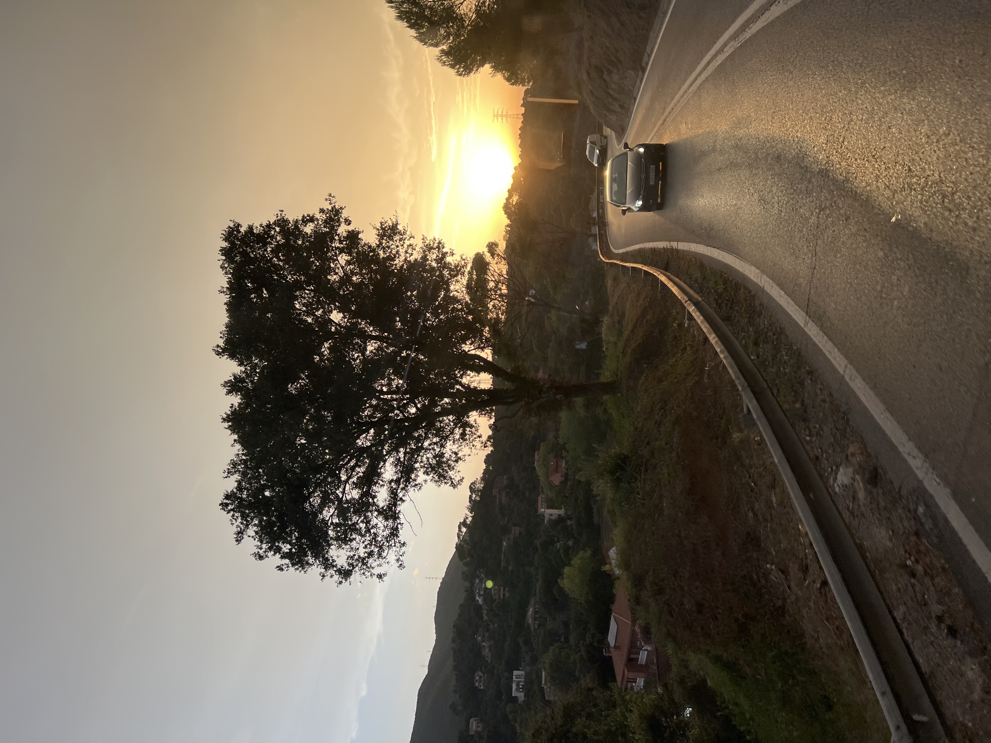 The image depicts a sunset on a winding road in Sant Fost de Campsentelles, Spain. The sun is setting behind a tree-covered hillside, casting a warm glow over the scene. A black car is driving on the road, its headlights on. The road is flanked by a guardrail and vegetation. In the background, houses are visible on the hillside, and power lines are also present. The sky is a gradient of orange and yellow, with some clouds. The overall impression is one of a peaceful evening drive through a rural landscape.