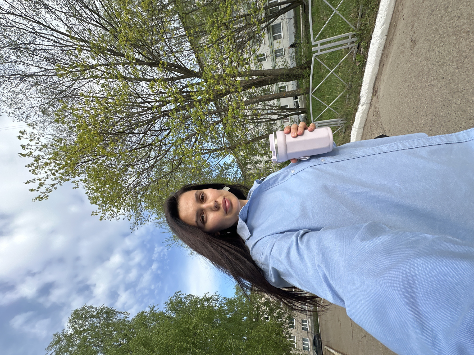 A young woman with long, dark hair is taking a selfie outdoors. She's wearing a light blue shirt and holding a light purple container in her right hand. The background shows trees with fresh green leaves, suggesting springtime. Behind the trees, there's a building with windows and a section of a white fence. The selfie seems to be taken on a walkway or path near a street. Given the prompt, the location is assumed to be in the city of Chaikovsky, Russia.