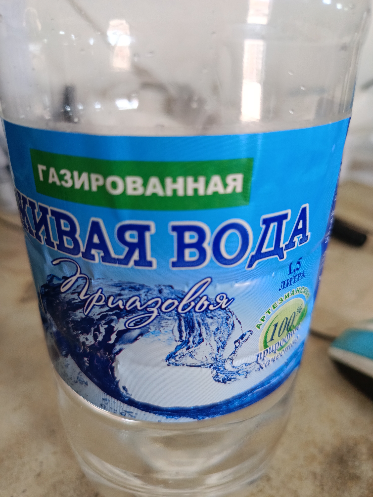 A close-up, eye-level, indoor shot shows the label of a clear plastic bottle of sparkling water. The label is primarily blue and green. The top portion of the label, in green, reads "ГАЗИРОВАННАЯ" (GAZIRUVA NNAYA), meaning "sparkling" in Russian. Below this, in large, white, stylized letters outlined in blue, are the words "ЖИВАЯ ВОДА" (ZHI VAYA VODA), meaning "living water". Below this, in a stylized blue script, are the words "Приазовя" (Priazovya), possibly referring to the Azov region. To the right of the script, in smaller blue text, is "1.5 ЛИТРА" (1.5 LITRA), indicating 1.5 liters. Further to the right, in a green oval with a white outline, is "АРТЕЗИАНСКА" (ARTESIANSKAYA), meaning "artesian". Inside the oval, in large green numbers, is "100%" (100 percent), with smaller text below reading "природное" (prirodnoye) and "качество" (kachestvo), meaning "natural quality". The water in the bottle appears clear, with small bubbles visible on the inside surface of the plastic. The bottle is positioned on a light-colored, textured surface, possibly a countertop, with hints of other objects in the background that are out of focus. The lighting is bright, with reflections visible on the plastic bottle. The scene is located indoors, in Berdiansk, Ukraine.