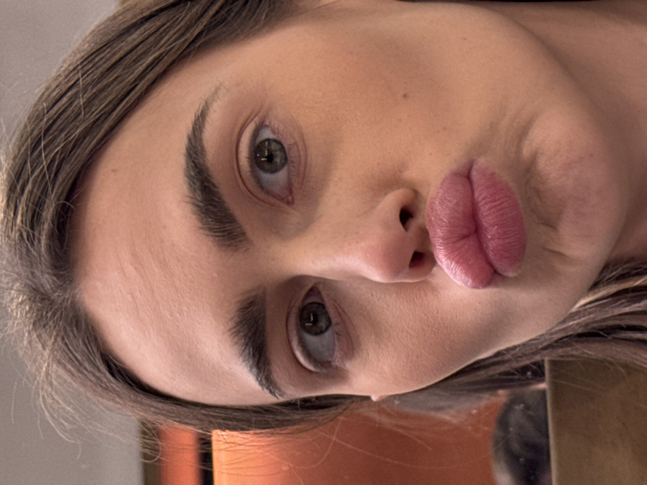 A close-up portrait depicts a single female subject facing the camera. She has brown hair, thick eyebrows, and light-colored eyes that gaze directly forward. Her lips are formed into a prominent pout. Her skin appears fair with some minor texture visible on the forehead and cheek. The background is out of focus, displaying indistinct warm orange-brown and darker tones on the left, with no discernible objects or features. The image provides no visual cues to determine its location in Verona, Italy, or any other specific geographical area.
