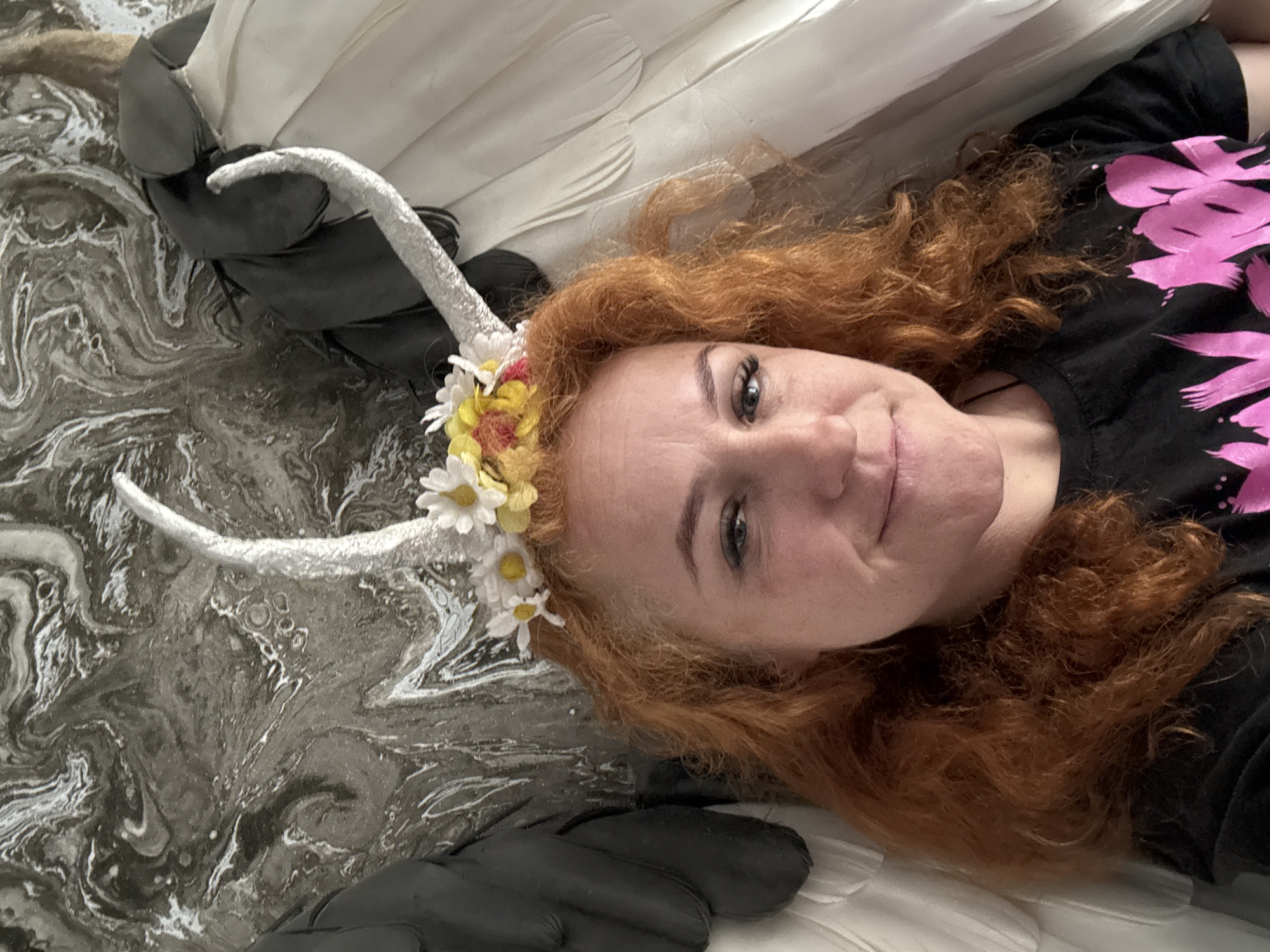 A woman with red curly hair is lying on what appears to be a bed, looking at the camera. She is wearing a black t-shirt with a pink floral design and a headpiece with white horns and a garland of flowers, including daisies and yellow flowers. The bed is covered with a patterned gray, white, and black material. There are also white and black elements around her, possibly wings or parts of a costume. It is not possible to verify the location being Chaikovsky, Russia with the provided image.