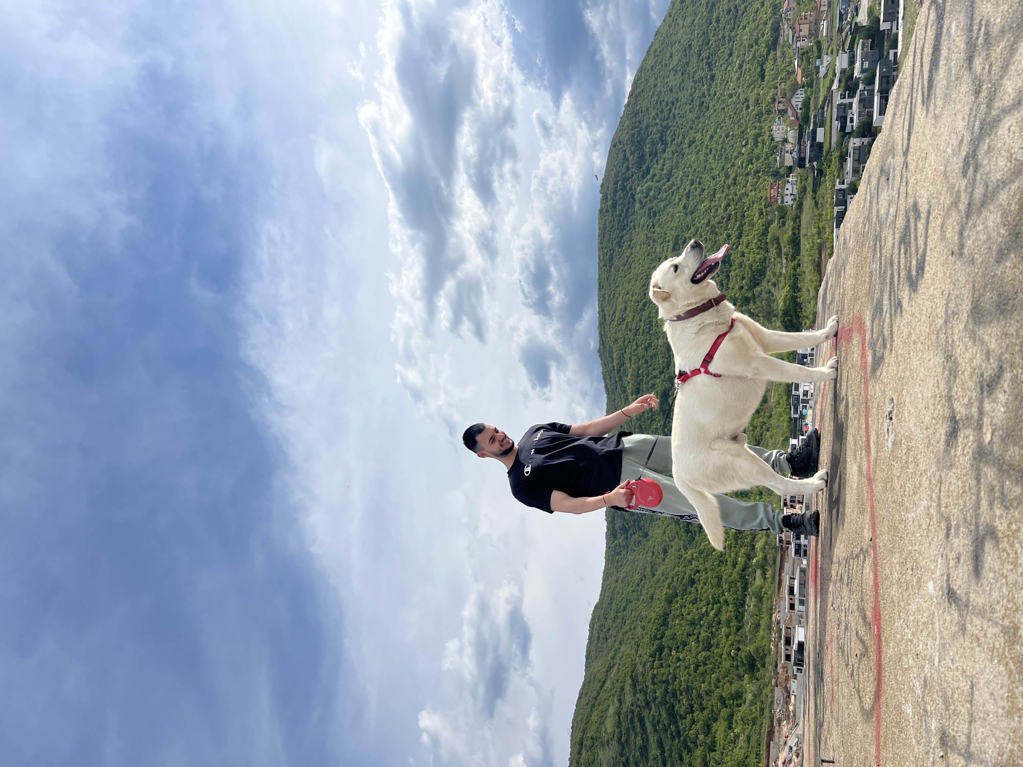 The image captures a man and a dog on a concrete structure, with a lush green hill and houses in the background. The man is wearing a black t-shirt, grey sweatpants, and black shoes. He is holding a red retractable leash. The dog, a large white breed, is standing on the concrete with its front paws on the edge. It has a red harness and collar, and its tongue is hanging out. The sky is visible with scattered clouds. This location is likely Markovo, Bulgaria.