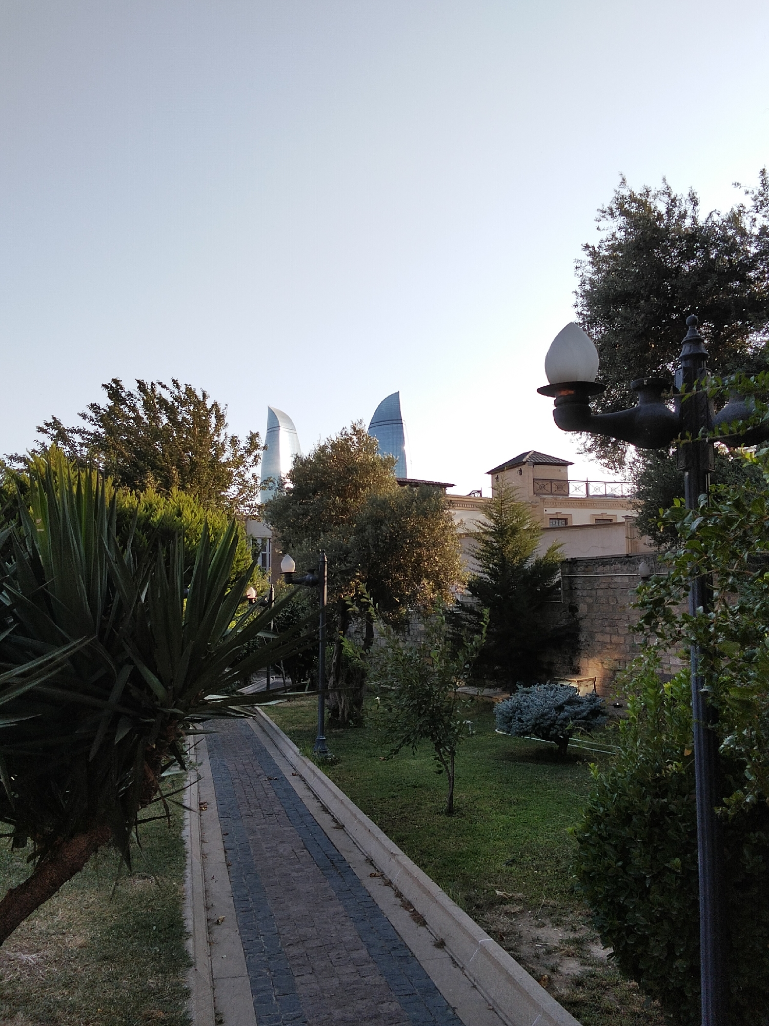 The image depicts a pathway in a park in Sabail Raion, Azerbaijan, leading towards some modern, flame-shaped skyscrapers in the distance. The pathway is paved with dark and light grey bricks, bordered by a concrete curb and grass. To the left of the pathway, a large, spiky green plant is prominent, with a tree trunk visible. Further down the pathway, a black lamppost with a white, bell-shaped light fixture stands to the right, partially obscuring a traditional stone building with a prominent upper section and a tiled roof. The background is dominated by lush green trees and the distinctive architecture of the Flame Towers. The sky is clear and pale blue.