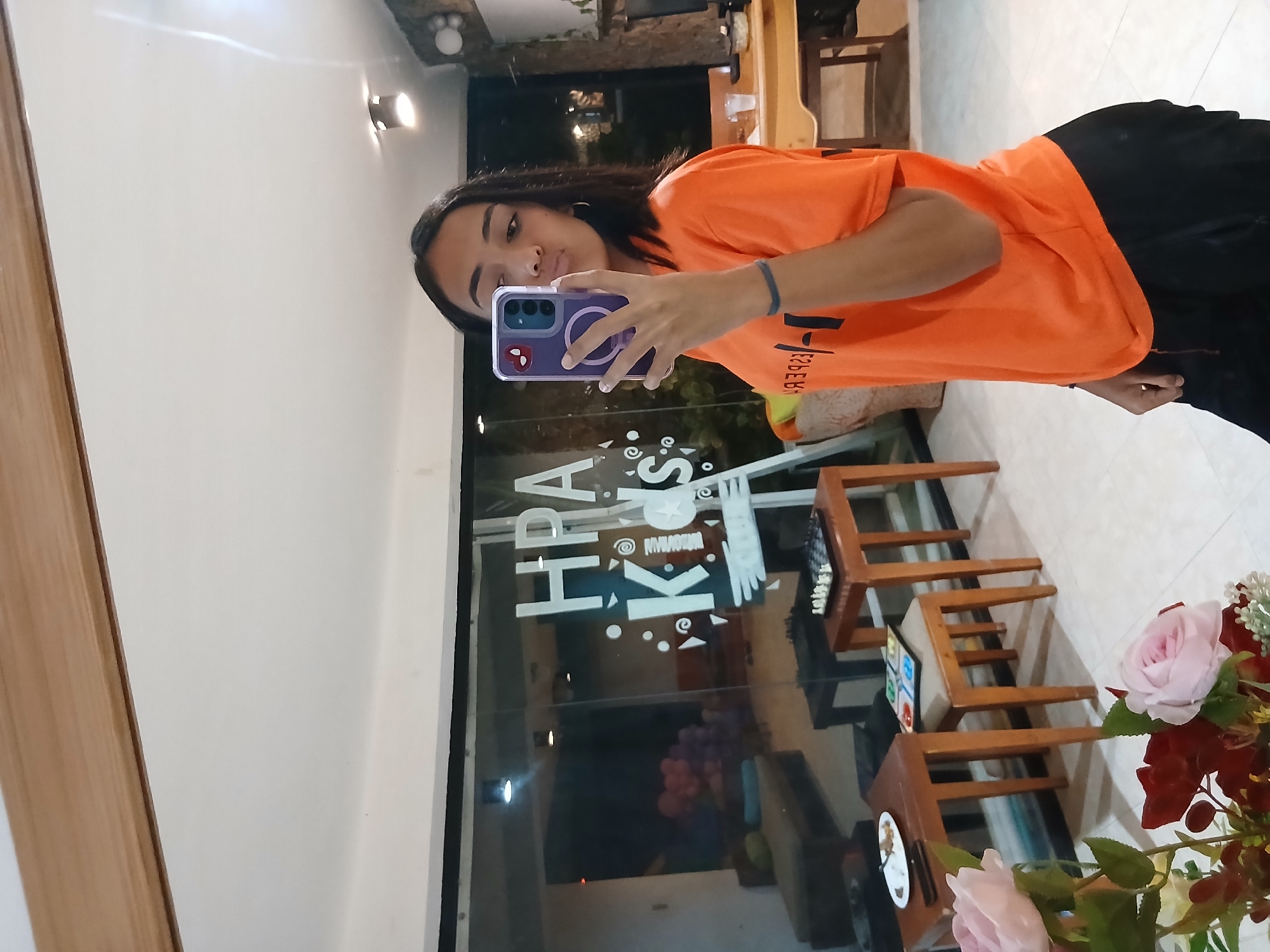 A person is taking a selfie in a reflective window. The person is wearing an orange shirt and black pants. The reflection in the window shows a room with chairs, a table, and a floral arrangement. The window has the text "HRA Kids" written on it. Based on the text and the style, the location is likely a children's educational center or playroom in Playa El Agua, Venezuela.