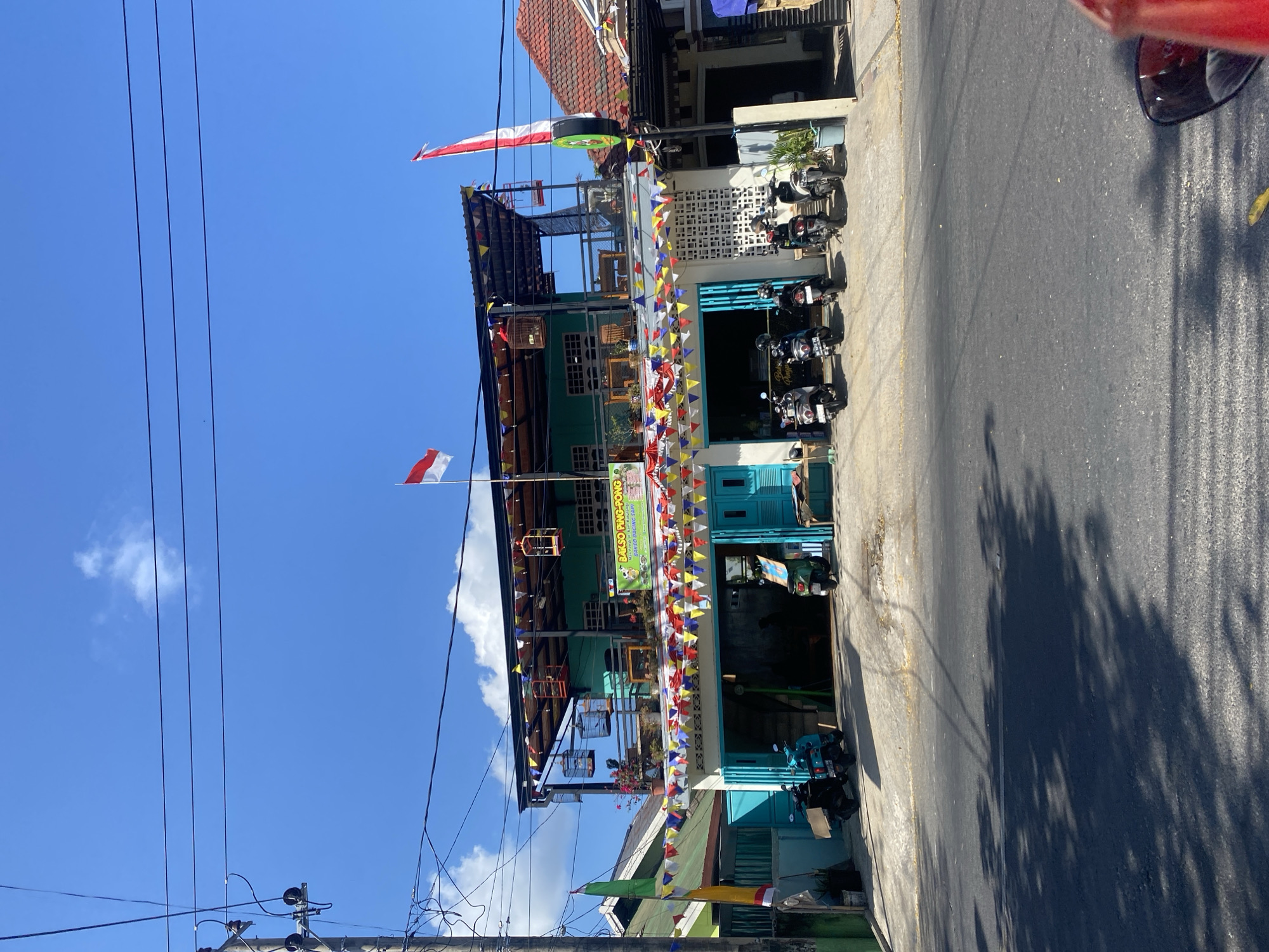 The image depicts a storefront in Bantaeng, Indonesia, on a sunny day with a clear blue sky. The building is a two-story structure painted in a vibrant turquoise color. The ground floor features a wide, open garage area with turquoise doors. Several motorcycles are parked in front of the garage and inside. A banner above the garage entrance reads "BALSO PING-PONG" and another smaller banner beneath it has text that appears to be in Indonesian.
The second floor has a balcony area with tables and chairs, accessible from the lower level via stairs that are not fully visible. The balcony is adorned with decorative flags in various colors, creating a festive atmosphere. Birdcages are suspended from the balcony's roof.
Two Indonesian flags, red and white, are prominently displayed; one flies from a pole on the roof of the building, and another is attached to a pole to the right of the building. Utility poles and electrical wires are visible against the sky. The street in front of the building is paved and shows shadows from trees. The overall impression is a bustling local establishment, likely a restaurant or cafe, decorated for a celebration.
