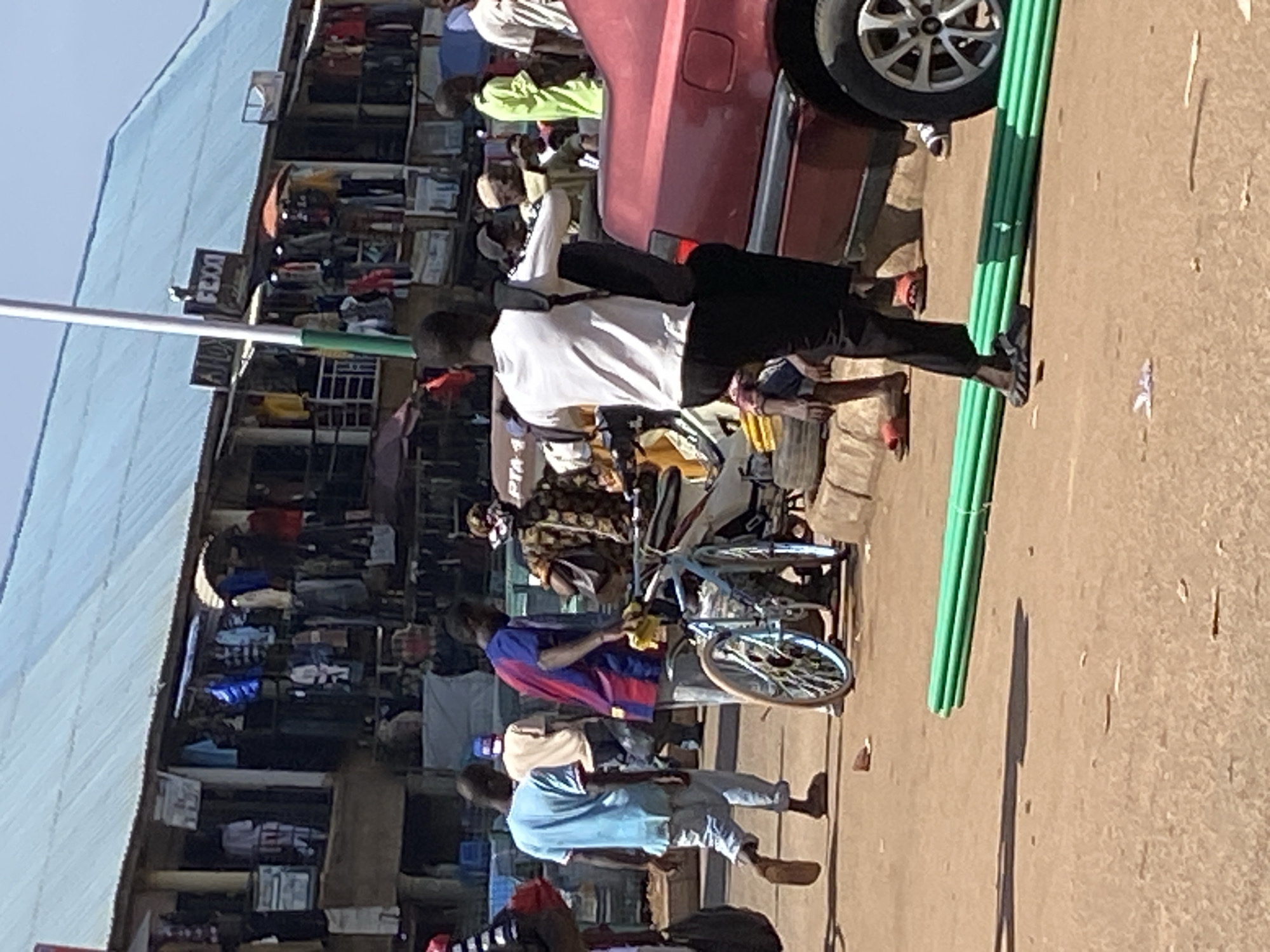 In Bukuru, Nigeria, a bustling marketplace scene unfolds under a blue corrugated metal roof. Several individuals are visible amidst stalls displaying clothing. In the foreground, a man in a white t-shirt and black pants carries a backpack, walking towards the right. To his left, another man in a red and blue striped shirt walks past a bicycle and a red car. Further in the background, a person in a light blue shirt walks away from the camera, and other figures are dispersed throughout the market. Green pipes are laid out on the ground in the foreground, partially obscured by shadows. The ground is dusty, typical of an outdoor market environment.
