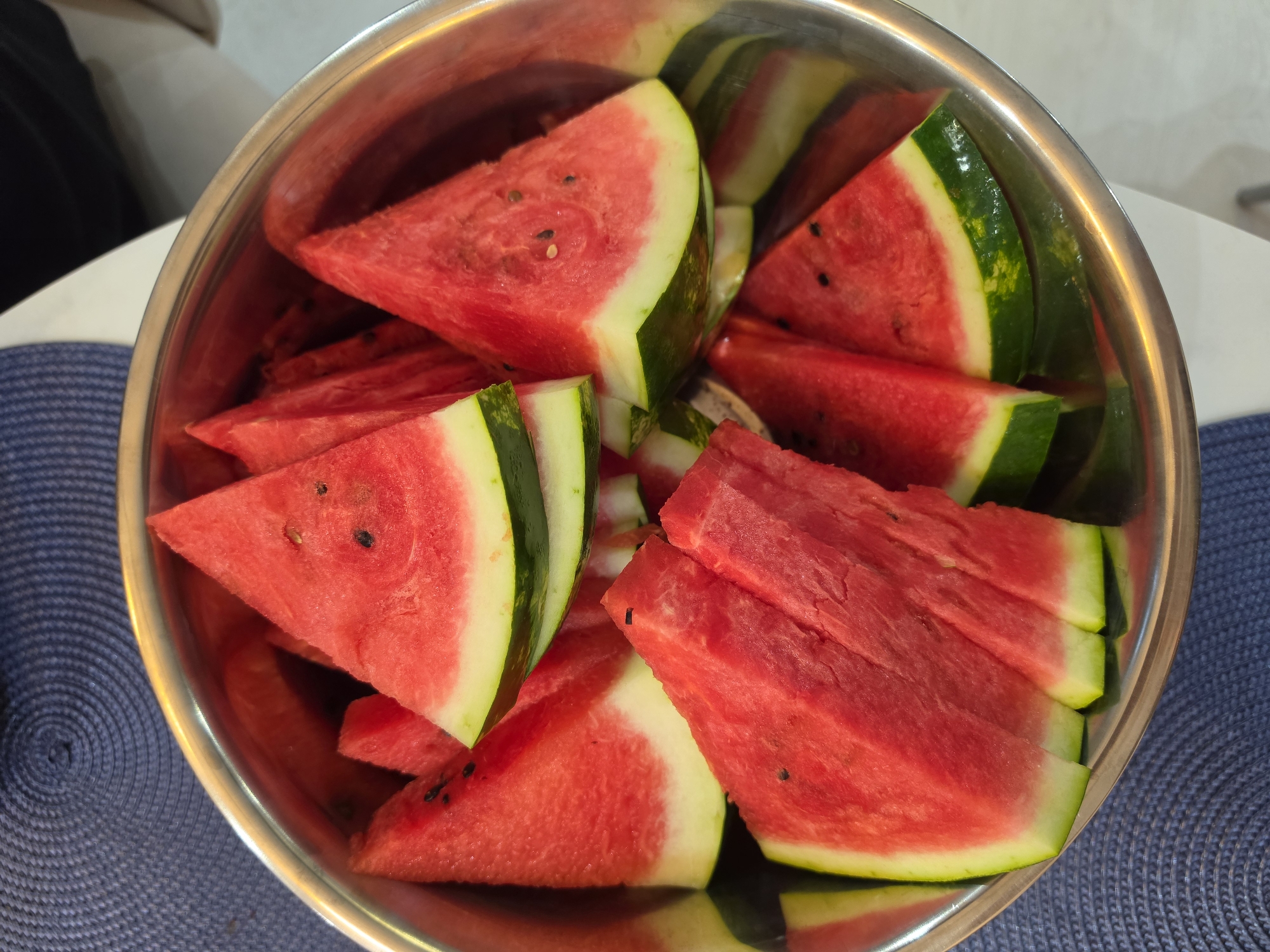 A close-up overhead shot shows a stainless steel bowl filled with sliced watermelon. The watermelon slices are triangular, with bright red flesh, white rind, and green skin. Some slices have black seeds visible. The bowl is placed on a blue, textured placemat, and a white surface is visible in the background. The scene appears to be indoors.