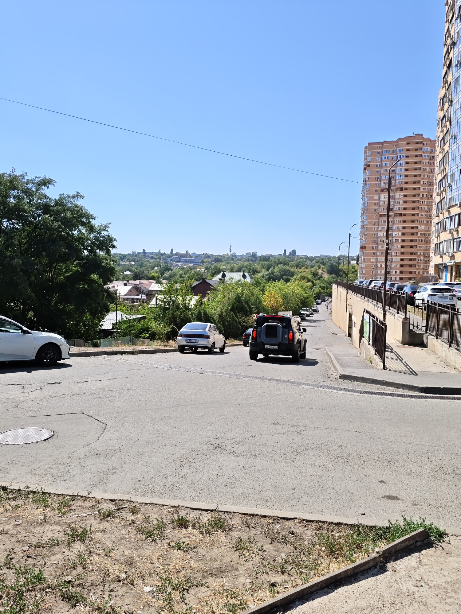 The image depicts a street scene in Rostov-on-Don, Russia, on a clear, sunny day. In the foreground, a light-colored sedan is parked on the left side of the road, facing away from the camera. Further down the street, a dark-colored SUV is turning, and another white sedan is parked on the right. The road is paved and appears to be in moderate condition, with some cracks visible.
In the mid-ground, a residential area with houses and trees stretches across a gentle slope. Beyond this, a modern, multi-story apartment building dominates the right side of the frame. This building features a facade of light brown bricks and numerous windows. A dark metal railing runs along a pathway next to the building. Several cars are parked in a row along this pathway.
The background shows a hazy, distant cityscape with buildings and trees under a bright blue sky. A thin black wire stretches horizontally across the upper portion of the image. The overall impression is a typical urban environment with a mix of older and newer architecture.