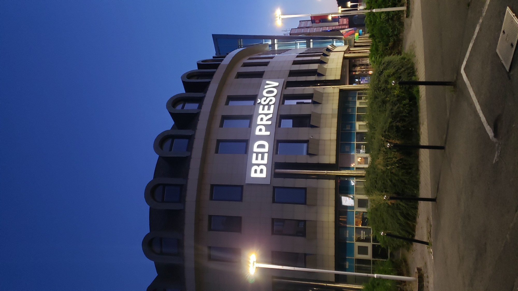 The image shows the building of BED PREŠOV, a business center located in Prešov, Slovakia, at dusk. The building is a modern, curved structure with multiple floors of windows. A prominent white sign on the facade reads "BED PREŠOV" in large, illuminated letters. The sky is a deep blue, indicating the late evening. In front of the building, there is a paved area with bollards and a dense green hedge. Streetlights illuminate the scene, casting a warm glow on the building and surrounding pavement. The overall impression is one of a well-lit and modern commercial space.