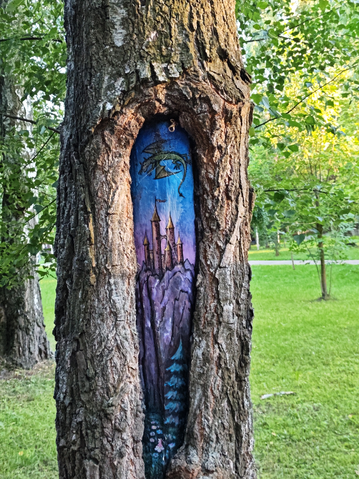 The image shows a close-up of a tree trunk with a painted mural inside a hollow. The mural depicts a fantasy scene with a castle perched on a rocky mountain, a dragon flying in a vibrant blue and purple sky. The tree is in a park-like setting with green grass and other trees visible in the background. The overall impression is of a whimsical artistic intervention in a natural environment. The location is specified as the city of Shcherbinka, Russia. There are no people or animals present in the image.