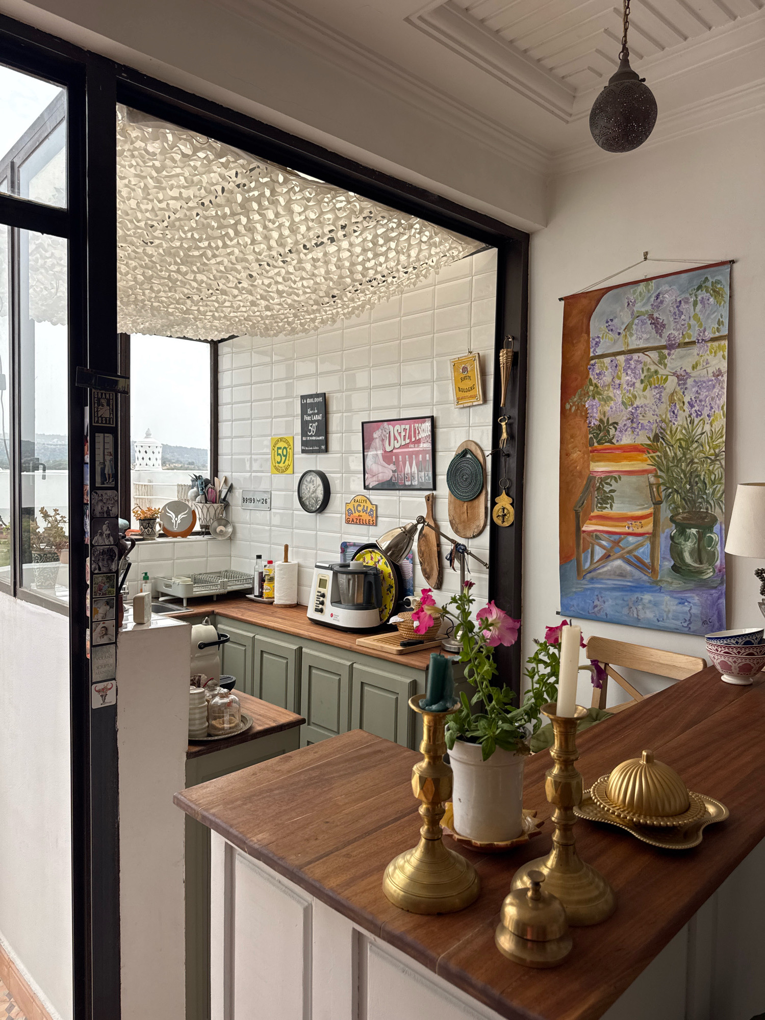 The image depicts an indoor scene, likely a kitchen or dining area, with an open doorway revealing an outdoor view. The indoor space features a wooden countertop in the foreground with brass candlesticks, a potted plant, and a golden cloche on a decorative tray. Behind the countertop, a kitchen area is visible with green cabinetry, a wooden countertop, a white tiled backsplash, various kitchen appliances including a food processor, a dish rack, and a roll of paper towels. Decorative items such as signs, a clock, and artwork adorn the walls.
The doorway opens to a sunlit area with white tiled walls and a patterned ceiling. Beyond this, a glimpse of the outdoors shows a distant landscape with hills and some structures, suggesting a location on the outskirts of a city. The style of the artwork and the overall decor hints at a North African aesthetic.
The scene is static, with no people present. The lighting is natural, coming from the open doorway and likely overhead fixtures. The overall impression is one of a lived-in, yet tidy, domestic space. The mention of Essaouira, Morocco, in the prompt suggests that this scene is likely situated in or near this coastal city.