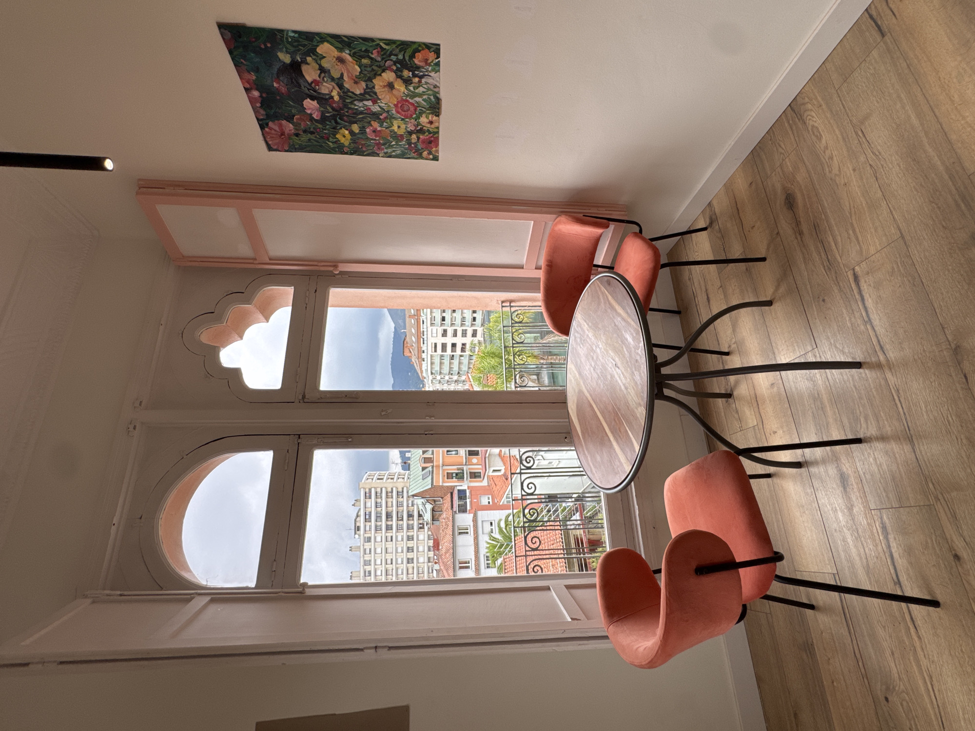 The image depicts a cozy seating area near a large arched window overlooking a city. The window frames a view of buildings with terracotta roofs, a lush green palm tree, and distant hills under a cloudy sky. Inside, two vibrant coral-colored chairs are placed around a round wooden table with a black metal base. The room features light-colored walls and a light wood floor. On the wall to the right, a colorful painting of flowers and a partially obscured face hangs. A black cylindrical spotlight is mounted on the ceiling. The window has white shutters, and part of a pink decorative wooden frame is visible on the right side of the window. The scene conveys a sense of calm and relaxation, perhaps in a cafe or a residential space. It is not possible to determine if the location is in Oviedo, Spain, from the image alone.