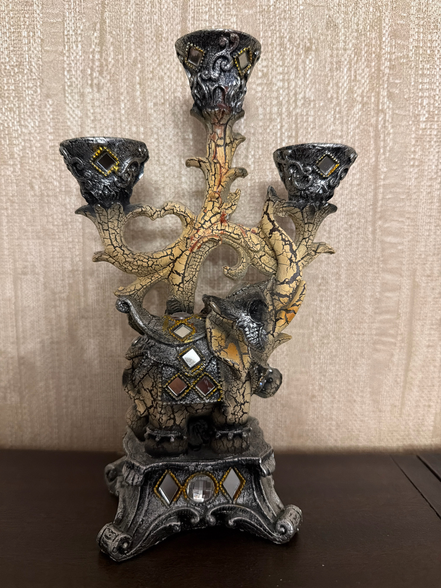 A detailed, front-facing, medium shot depicts an ornate, multi-branched candelabra designed to resemble a tree with an elephant at its base. The candelabra is made of a dark, possibly metallic material, with distressed crackled paint effects in shades of yellow and orange on the tree-like branches. Three candle holders, each with decorative filigree and small diamond-shaped mirrored insets with gold trim, are positioned at the top of the structure. The elephant at the base of the candelabra is depicted with intricate detailing, including textured skin, and is adorned with diamond-shaped mirrored insets. The candelabra rests on a decorative, dark-colored base that also features mirrored insets. The background is a textured, neutral-colored wall. The scene is lit evenly, and the object is centered within the frame. This object is located inside a residential property in Sabunchu Raion, Azerbaijan.