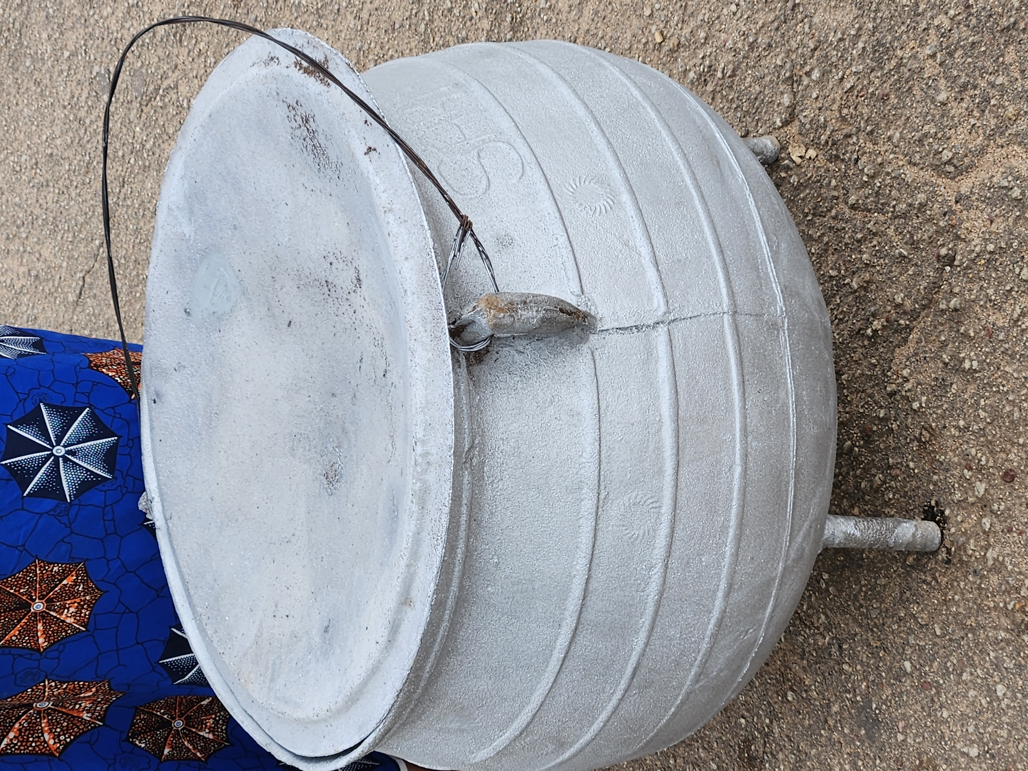 The image shows a large, silver-colored pot with a lid, lying on its side on a textured, gravelly surface. The pot has a round, bulbous body with horizontal ribbed detailing. Two short, cylindrical legs extend from the side of the pot. A thin, black wire handle is attached to the rim of the pot and extends upwards and over the lid. A small, weathered object, possibly a piece of stone or metal, is attached to the wire handle near the lid.
The lid of the pot is also silver and appears to be slightly dusty or weathered. It fits snugly onto the pot. To the left of the pot, a portion of fabric with a blue background and a repeating pattern of umbrellas in orange and black is visible. The umbrellas are detailed with white accents.
The scene appears to be outdoors, with the textured ground suggesting a paved area or compacted dirt. The lighting suggests it could be daytime, but there are no strong shadows to indicate a specific time. There are no people visible in the image, only the pot and the fabric. The overall impression is that of a utilitarian object, perhaps used for cooking or storage, placed in an outdoor setting. There is some faint text or markings on the side of the pot, including what appears to be a stylized "S" and a circular emblem with radiating lines.