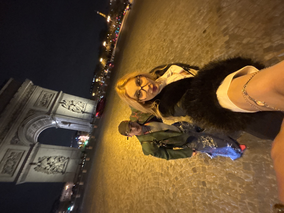 An outdoor nighttime photograph depicts two individuals posing on cobblestone pavement in Paris, France. The prominent landmark, the Arc de Triomphe, is illuminated in the background, slightly tilted to the left due to the wide-angle perspective. The Eiffel Tower is faintly visible, illuminated, in the far distance to the right.
The foreground is occupied by a female subject, partially visible as the camera holder. She has blonde hair and is wearing eyeglasses, a white collared shirt, and a dark coat with a fur collar. A silver-colored chain accessory is visible on her right arm or wrist. Her lips are slightly parted.
Behind her, slightly to the left, stands a male subject. He wears a dark cap, a light-colored inner garment, a dark green jacket, and dark pants that appear distressed or torn. He looks directly towards the camera with a neutral expression.
The ground is uniformly paved with cobblestones, illuminated by a warm, yellowish artificial light source, casting strong reflections on the surface. In the mid-ground, a red vehicle is visible on a road behind the subjects. Distant city lights and other vehicle lights delineate a busy urban environment beyond the Arc de Triomphe. The overall sky is dark, indicating night.