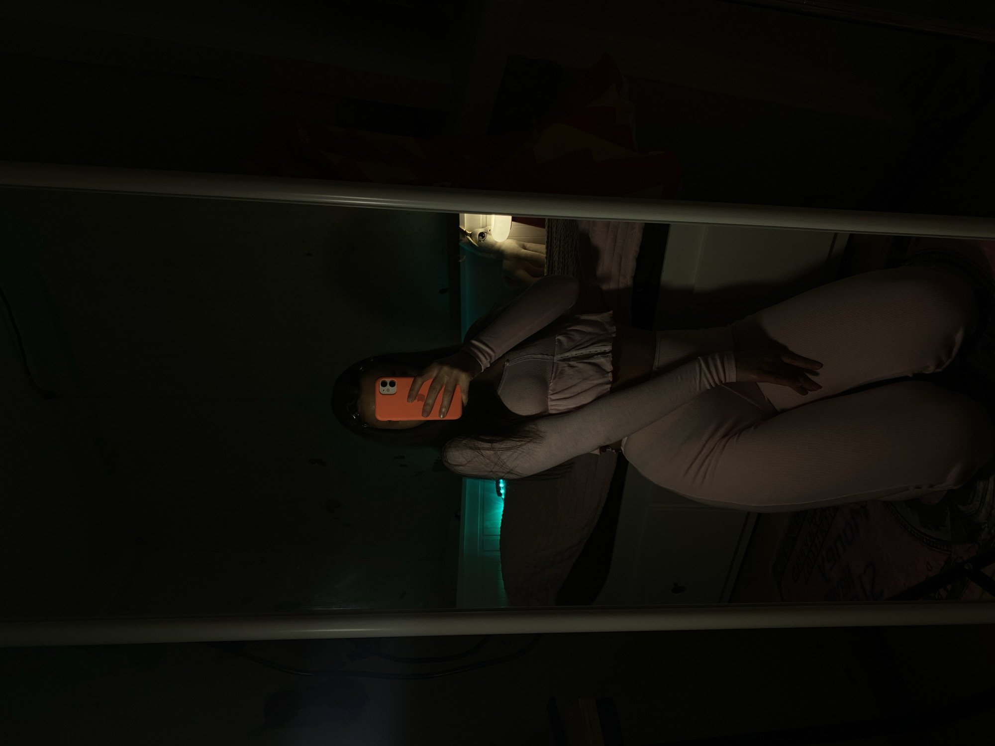 A person in a pink outfit holds an orange smartphone, capturing a selfie-style shot in a dimly lit space. The person appears to be lying down, with their legs and torso visible. A white bar runs across the foreground, possibly a bed frame or other structural element. Behind the person, various indistinct objects can be seen. The overall environment is dark. There is no indication of the city of Votkinsk, Russia, in the image.