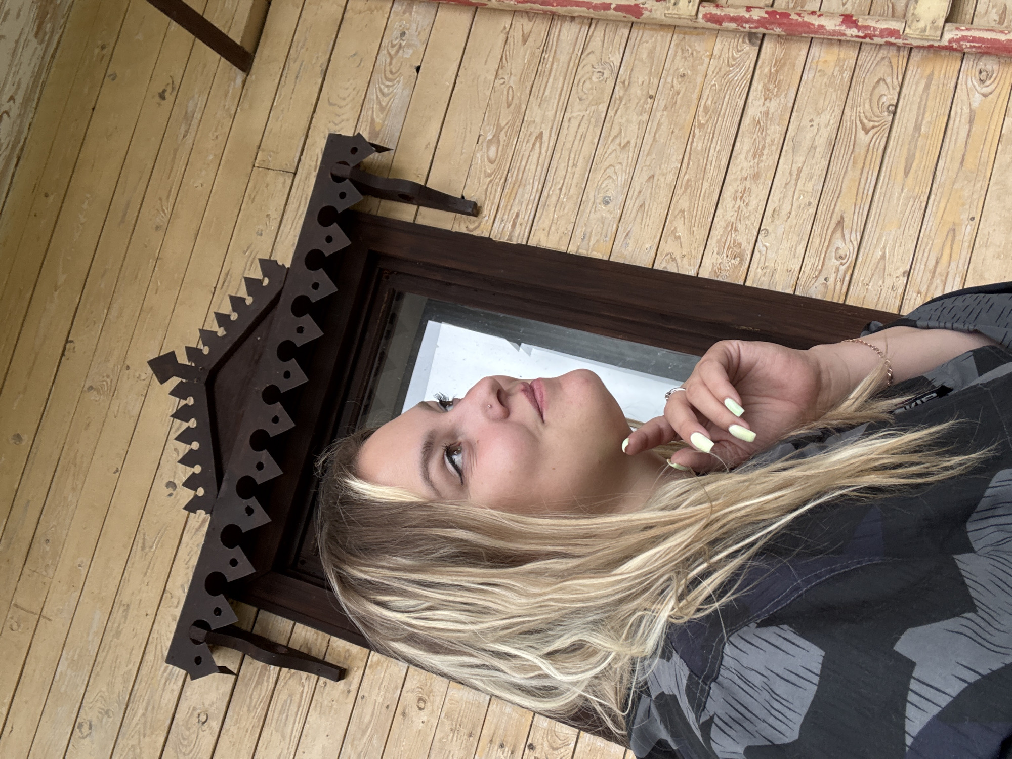 The image depicts a young woman with blonde hair, wearing a grey and black camouflage jacket, standing in front of a wooden building. She is looking upwards and to the right, with her right hand touching her chin. A portion of a window frame with decorative woodwork is visible behind her. The building's exterior is constructed of horizontally laid wooden planks, painted a light yellow. The scene is set in Zavod Mikhailovskii, Russia.