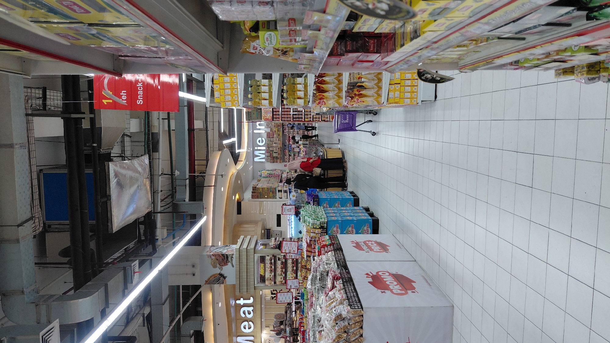 The image shows the interior of a brightly lit supermarket aisle in Purwokerto, Indonesia. The aisle is lined with shelves stocked with various food products, including packaged snacks, cooking oil, and instant noodles. On the left side of the aisle, large white displays with red lettering indicate "PROMO" and "SPECIAL PRICE." In the background, signs for "Meat" and "Mie Instant" (instant noodles) are visible. Several shoppers, including two women and a man, are seen browsing the shelves. A purple shopping cart is parked in the aisle. The floor is tiled with white square tiles. The ceiling features exposed ductwork and lighting fixtures.
