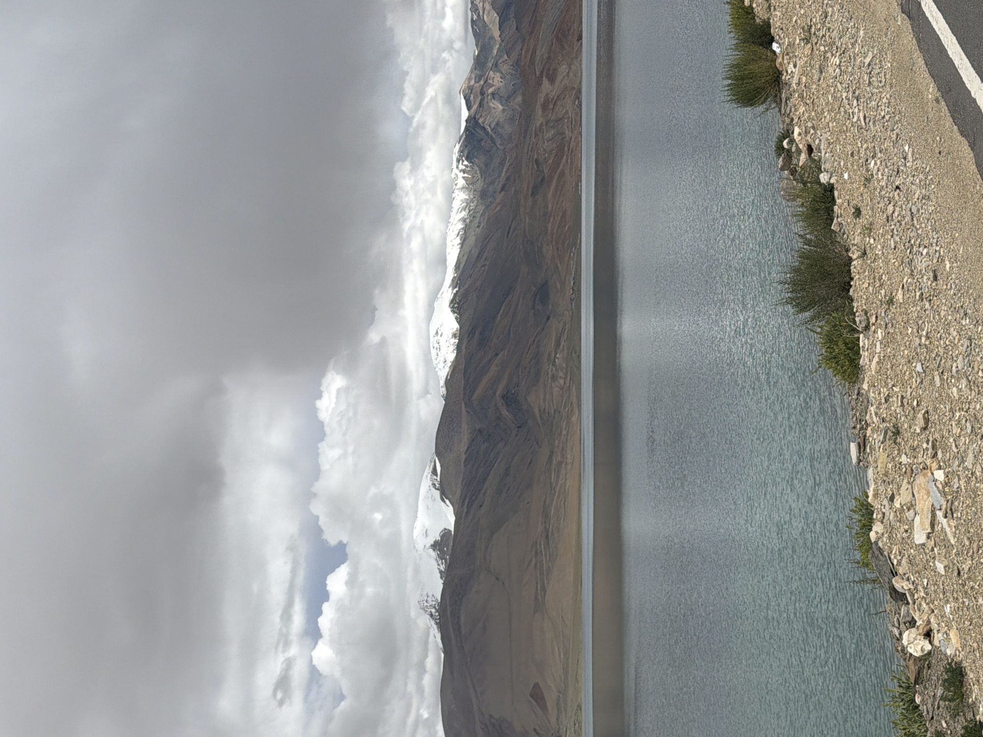 A wide shot captures a serene, serene lake reflecting the overcast sky. The lake's surface is calm, with gentle ripples distorting the reflection of the barren, mountainous landscape that borders it. The mountains are layered with shades of brown and tan, their peaks dusted with snow. The sky above is filled with heavy, grey clouds, obscuring any direct sunlight. The foreground of the image shows a rocky, dry terrain leading to the lake, with sparse, green bushes dotting the landscape. The road is visible on the far right of the frame. This is likely the location in Karzok, India.
