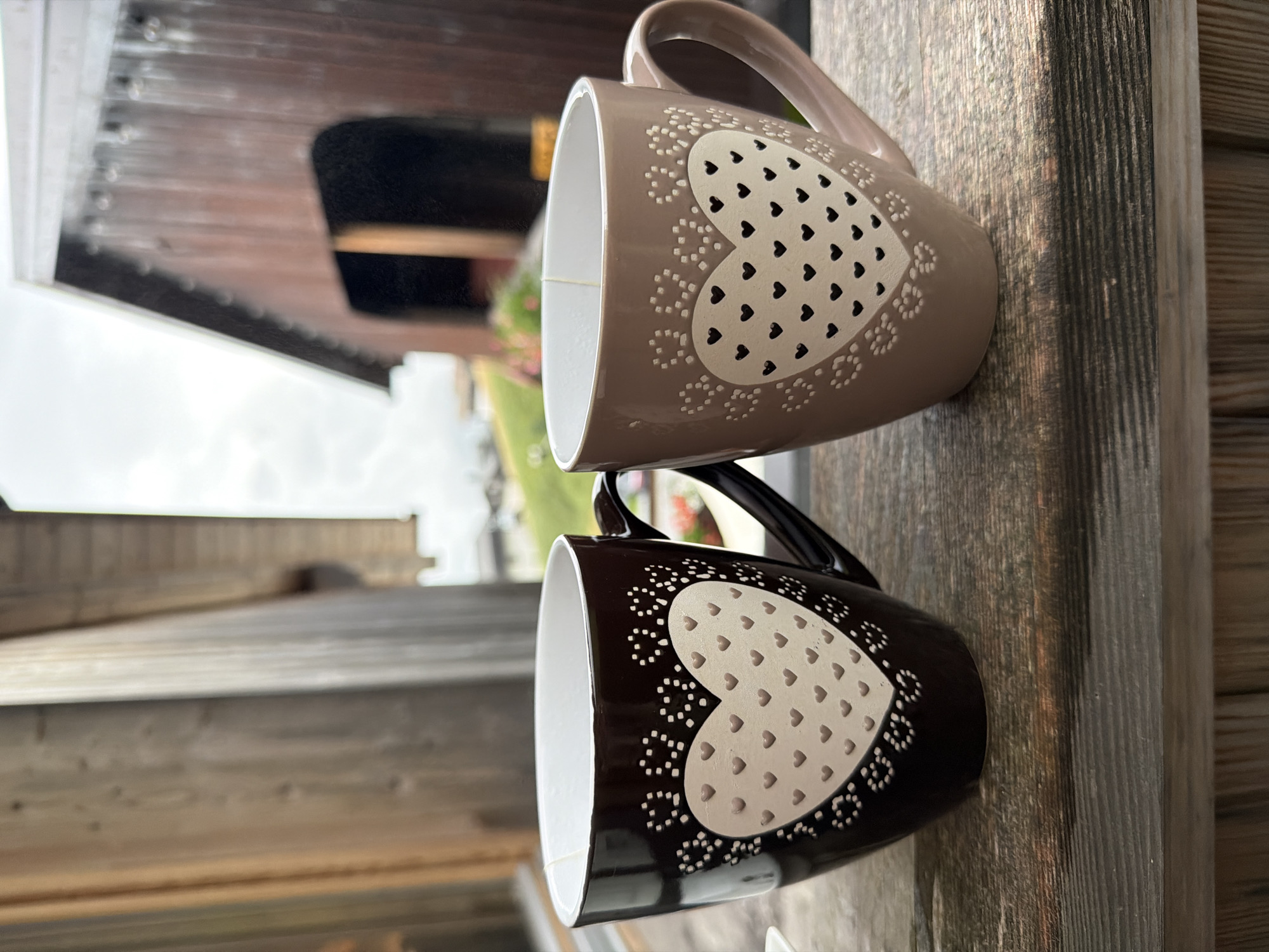 Two ceramic mugs with heart designs sit on a wooden surface. The mug on the left is dark brown with a light pink heart filled with small dark hearts. The mug on the right is light brown with a light pink heart filled with small dark hearts. Both mugs have a decorative border of dots and small hearts around the main heart design. In the background, a rustic wooden building with a dark brown roof is visible, along with greenery and a cloudy sky. The setting appears to be outdoors, possibly on a porch or patio.