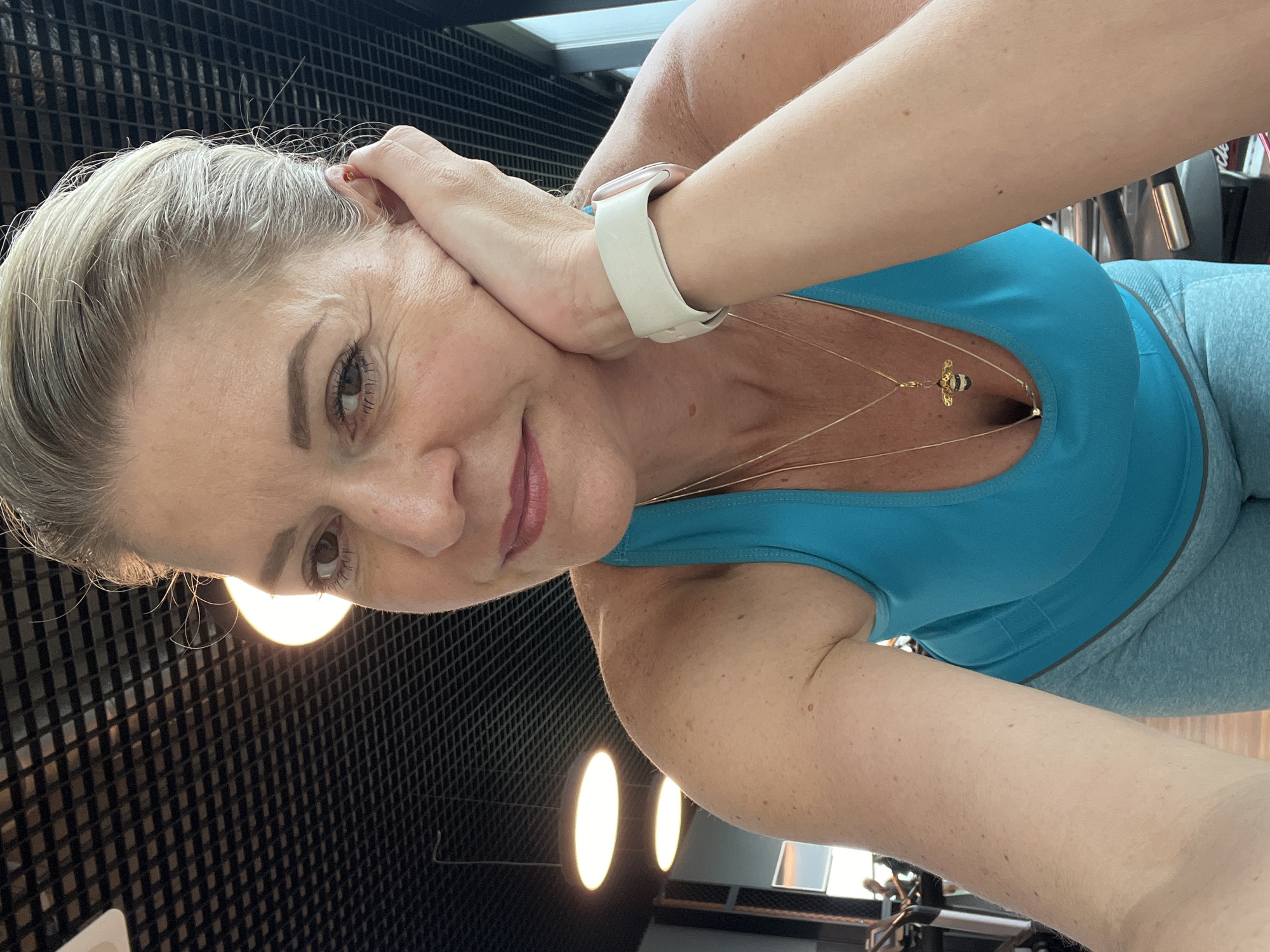 A woman with blonde hair pulled back is taking a selfie in what appears to be a gym. She is wearing a teal tank top and grey leggings. She has a white smartwatch on her left wrist and is wearing a necklace with a bee pendant. Her left hand is resting on her cheek. The background shows a black, textured ceiling with oval lights and a window with a view of gym equipment. There is no indication that this location is in Kaliningrad, Russia.