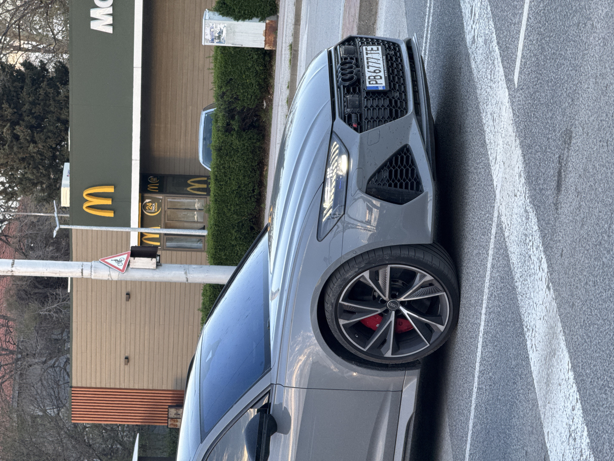 The image depicts a gray Audi RS7 parked on a crosswalk in front of a McDonald's restaurant. The license plate of the car reads "PB6777TE", suggesting the image was taken in Bulgaria. A traffic sign indicating a bicycle crossing is also visible near the car. The location is likely in the city of Plovdiv, Bulgaria, given the car's license plate and the presence of a McDonald's. The time of day appears to be daytime. There are no people visible in the scene.