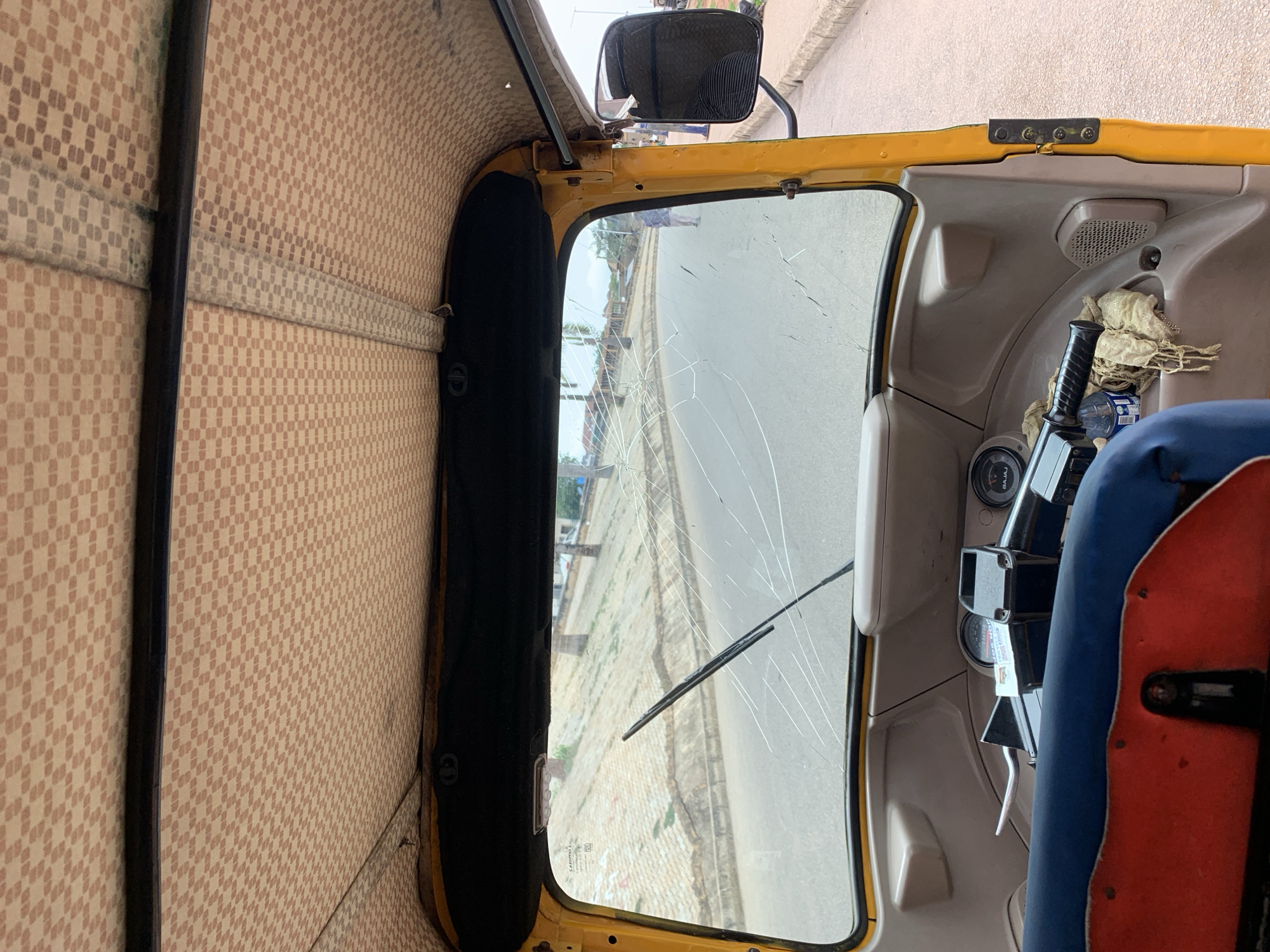 The image depicts the interior of a yellow autorickshaw, likely a Bajaj model, in an urban environment. The vehicle's cracked windshield reveals a view of a street or road. The interior features patterned upholstery and a steering column with gauges and controls. A portion of the driver's seat, covered in blue and red material, is visible. The exterior view suggests a street scene with buildings and pavement. The location is possibly in Kabong, Nigeria, but there are no specific visual elements to confirm the city's location directly.