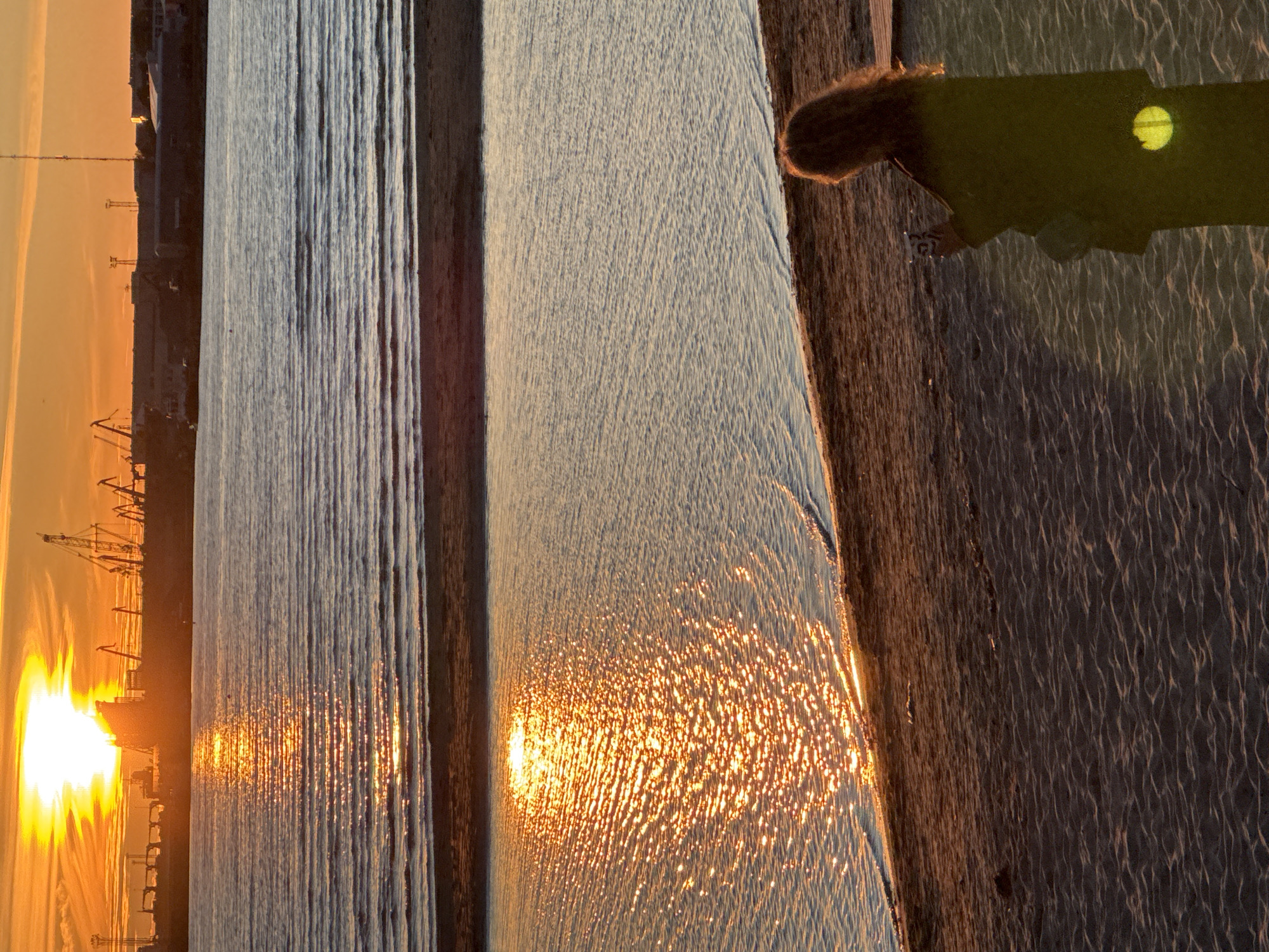 The image captures a serene sunset over the Baltic Sea in Tallinn, Estonia, as seen from the Pirita beach promenade. The sky is ablaze with warm hues of orange and yellow, with the sun partially obscured by clouds. The water reflects the vibrant colors of the sky, creating a shimmering pathway of light. In the distance, the silhouette of the Tallinn harbor is visible, with cranes and ships dotting the horizon. A lone figure, a woman with long, curly hair, stands on the sandy shore, holding a phone and seemingly taking a picture of the picturesque scene. She is dressed in dark clothing, with a light-colored bag slung over her shoulder. The sand on the beach exhibits a textured pattern, with subtle ripples from the receding tide. The overall impression is one of tranquility and natural beauty, enhanced by the warm glow of the setting sun.
