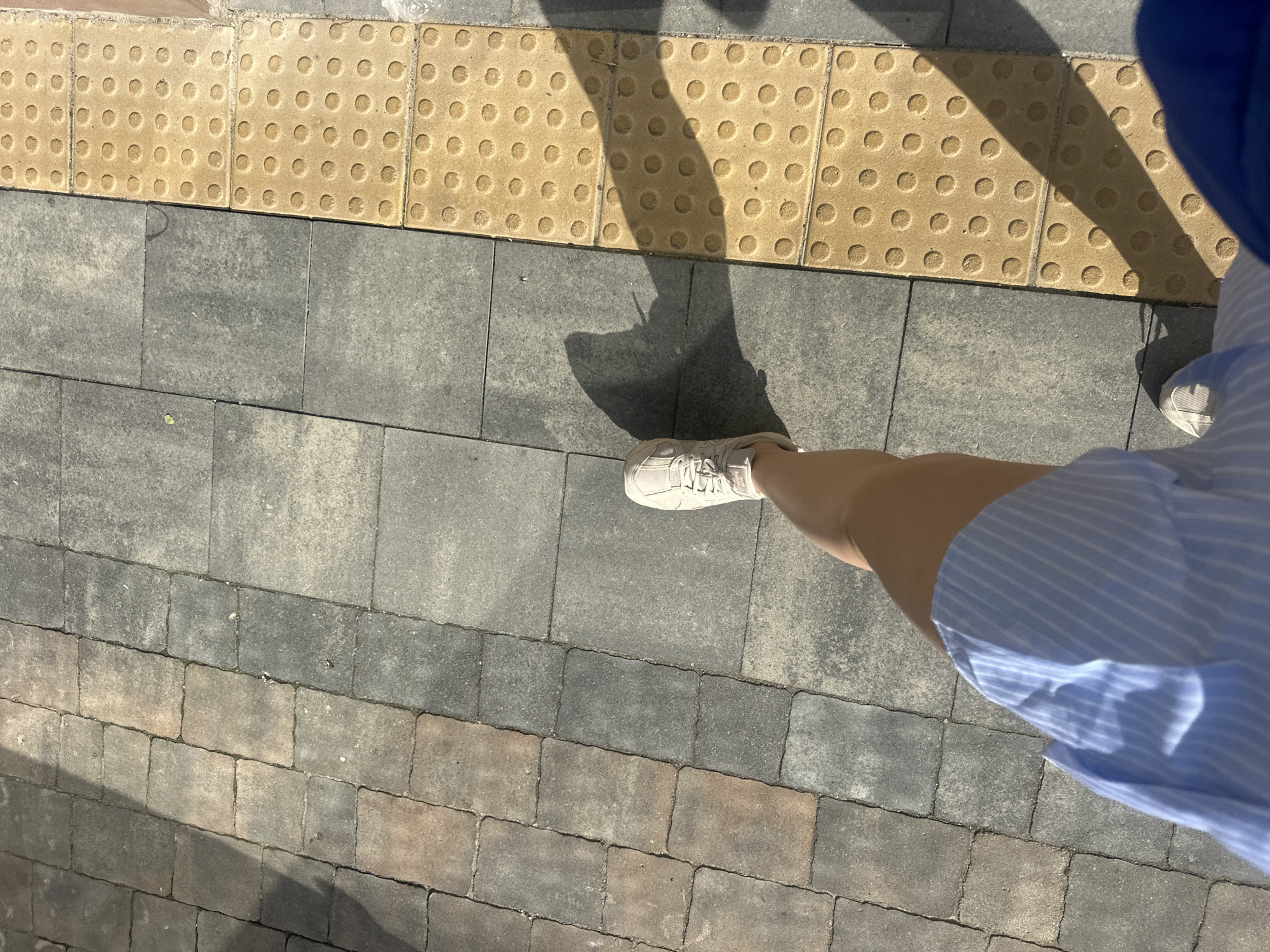 An overhead shot captures a person's legs and feet walking on a sidewalk composed of alternating gray and brownish rectangular paving stones. To the right of the paving stones is a row of yellow tactile paving tiles with raised circles, a common feature in urban environments for visually impaired pedestrians. The person is wearing light-colored sneakers and a blue and white striped dress. The sunlight casts a distinct shadow of the person and their leg onto the sidewalk. The scene appears to be a typical urban street or pedestrian walkway, possibly in Ternopil, Ukraine, given the context provided.