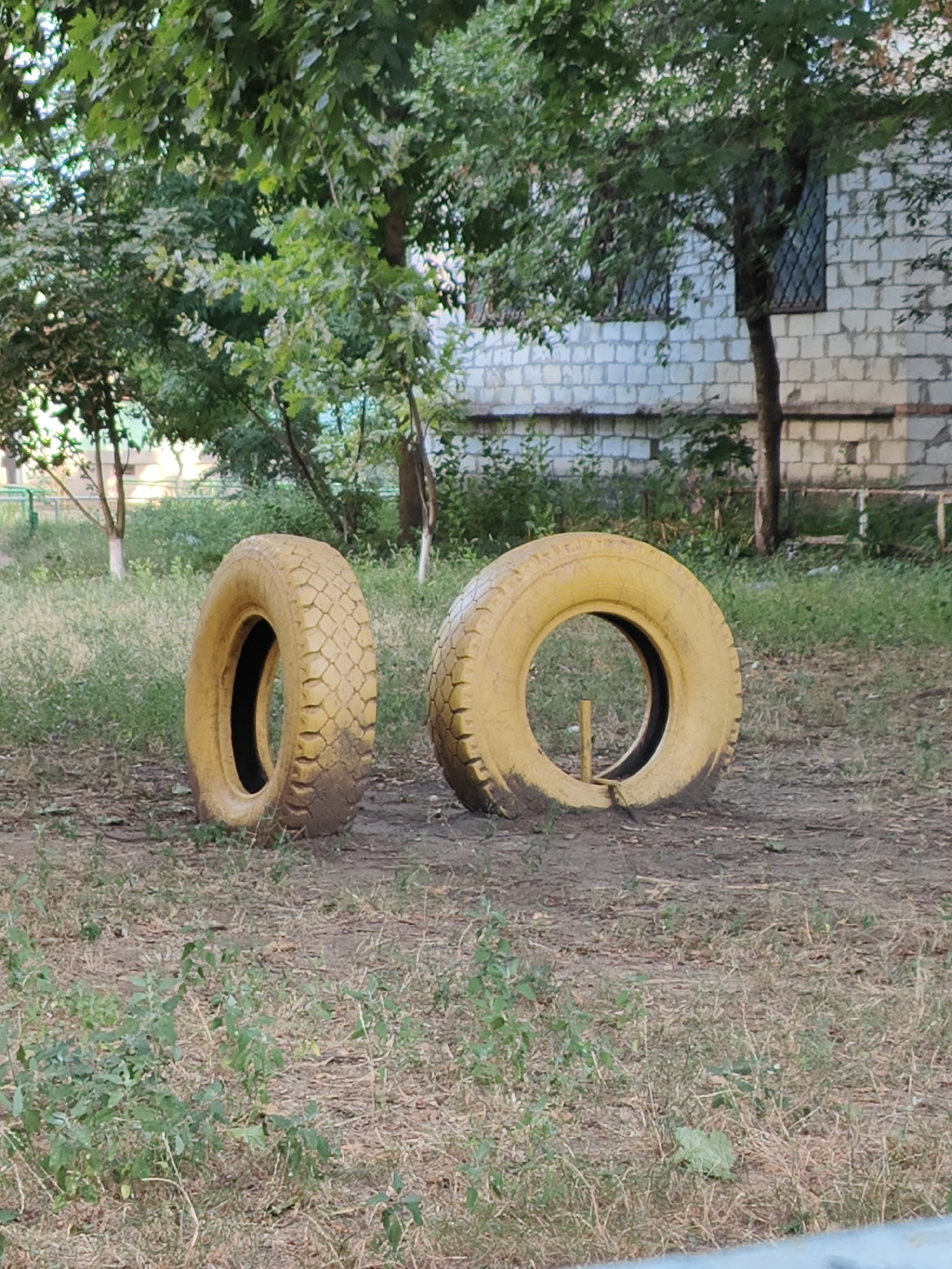 Two yellow tires, likely from a vehicle, are placed upright on a patch of dry grass and dirt. The tire on the left is positioned at a slight angle, while the tire on the right is more upright and has a thin, light-colored rod inserted through its center. In the background, lush green trees with dense foliage are visible, with a white brick building and windows with bars behind them. The scene appears to be an outdoor area, possibly a park or playground, in Tiraspol, Moldova. The lighting suggests it is daytime.