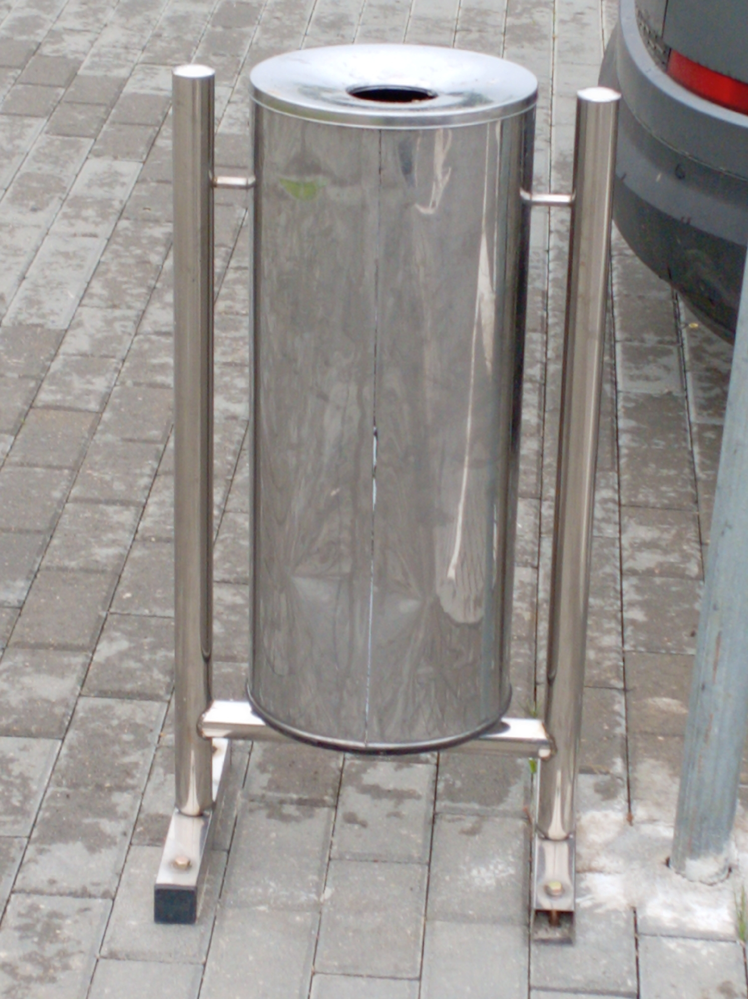A medium-shot displays a cylindrical, stainless-steel trash receptacle held by two sets of matching, upright frames. The receptacle features a circular opening on its top. The frames, also made of stainless steel, comprise vertical bars connected at the top and bottom to horizontal bars that grasp the container. At the base of each frame are rectangular feet with dark, non-slip pads. The trash can is positioned on a paved surface composed of rectangular, gray bricks. To the right, a portion of a gray vehicle with a red tail light is visible, alongside a cylindrical post set into the ground. The scene appears to be located in an urban or public area.