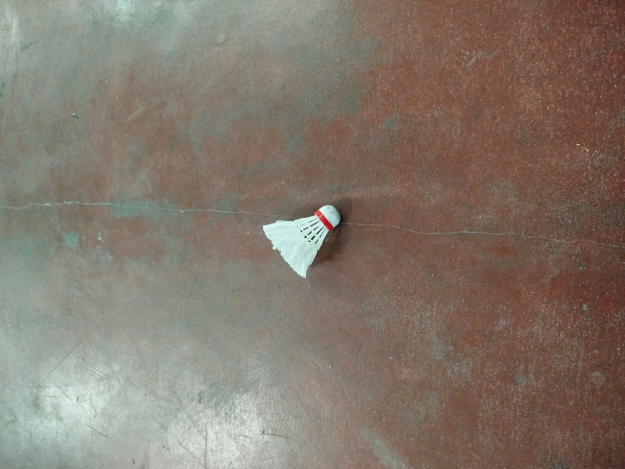 A single white and red badminton shuttlecock rests on a reddish-brown, worn, and scuffed concrete floor. The shuttlecock is positioned slightly off-center to the right and near the bottom of the frame, with its feathers fanned out. A thin, light-colored crack runs horizontally across the floor, passing just below the shuttlecock. The lighting suggests an indoor environment with some ambient light creating slight variations in the floor's color and texture. The scene is devoid of any human presence or activity. The location is identified as the city of Semin, Indonesia.