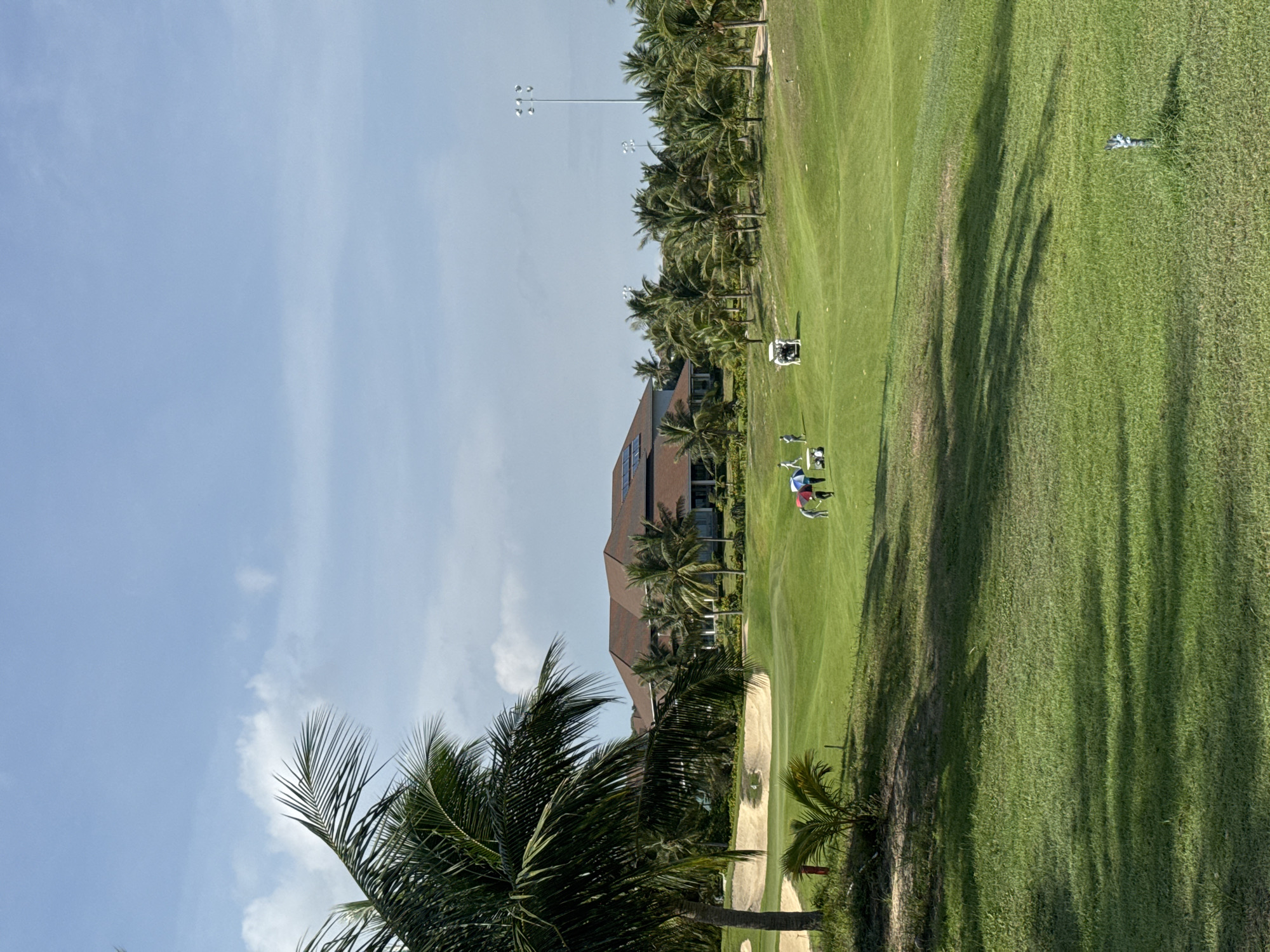 The image depicts a golf course with lush green fairways and several sand traps, set against a backdrop of palm trees and a large, brown-roofed building. The sky is blue with scattered white clouds. In the mid-ground, a group of people, some holding umbrellas, are visible on the course, along with golf carts. The scene suggests a warm, sunny day suitable for playing golf. The location is identified as Phú Thủy, Vietnam.