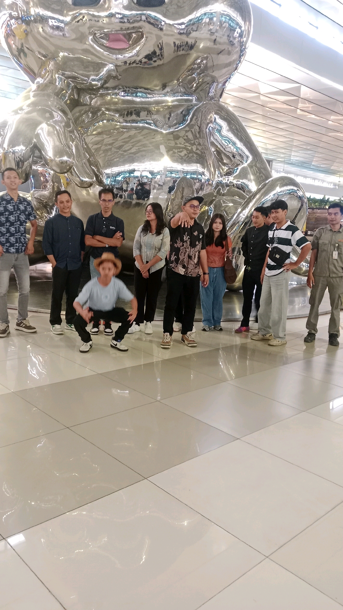 In a bright, indoor space with a polished tiled floor reflecting the overhead lights, a group of ten people stands near a large, shiny, silver sculpture of a cartoon character. The sculpture is the dominant feature in the background, its reflective surface distorting the surroundings. In the foreground, a young person wearing a straw hat and a light blue shirt is crouched down, appearing to pose. The other nine individuals are standing in a line behind this person, mostly facing the camera. The group consists of men and women of various ages, dressed in casual and semi-casual attire. The overall impression is a snapshot of a group posing for a photo in a modern public space, possibly an airport or a shopping mall, in Pajang, Indonesia.
