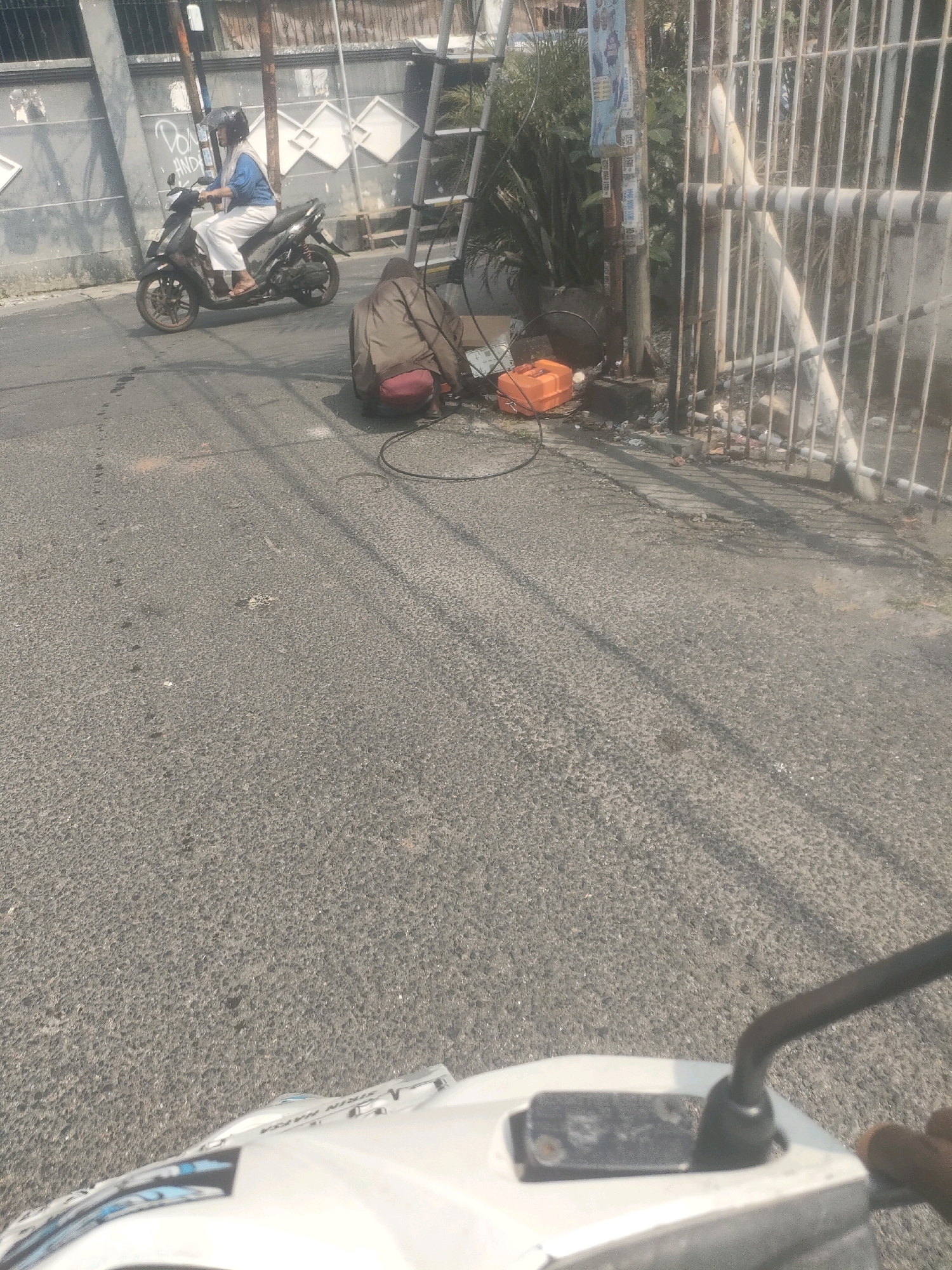 A person is riding a dark-colored scooter with a black helmet and blue shirt on a paved road. They are wearing white pants and sandals. To the right of the scooter, a person in a brown jacket and red shorts is crouched down on the road, working with some orange and black objects, possibly electrical equipment, and cables. Behind them is a white metal fence and some greenery. A ladder is also visible leaning against a pole. The scene appears to be in an urban environment in Surabaya, Indonesia, suggested by the context.