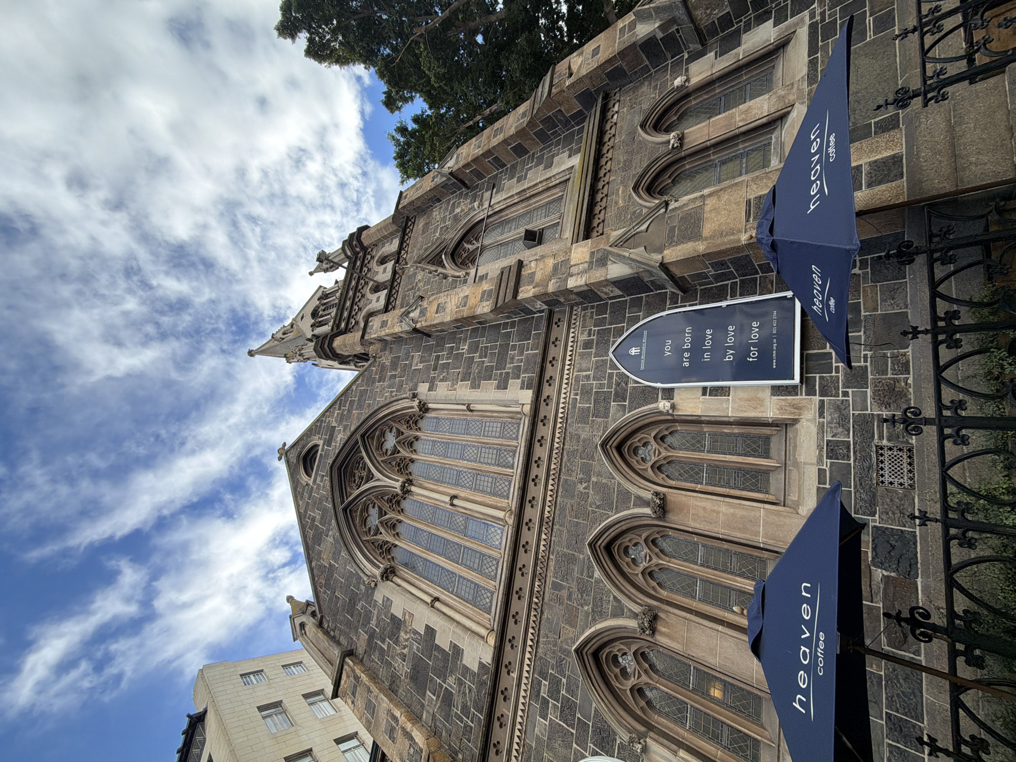 The image captures a detailed, low-angle view of a historic stone building, identified as the Central Methodist Mission in Cape Town, South Africa. The architecture is Gothic Revival, characterized by its imposing size, dark, variegated stone blocks, and intricate details such as large arched windows with delicate tracery and decorative buttresses. A tall spire or pinnacle rises towards the top of the structure, pointing towards a bright blue sky dotted with scattered white clouds, indicating a clear daytime setting.
In the foreground and to the right, two dark blue awnings or umbrellas are extended from the building's facade, each prominently displaying the text "heaven coffee" in white lettering, suggesting the presence of a café. Below one of these awnings, a rectangular white sign is affixed to the stone wall. The sign features a logo at the top left, representing the Central Methodist Mission. Below the logo, a spiritual message reads: "you are born in love by love for love." Further down, contact information includes the website "www.cmm.org.za" and the phone number "021 422 2744," the latter being a Cape Town area code that confirms the location.
To the bottom left, a portion of a more modern, lighter-colored building with multiple windows is visible, providing a stark contrast to the ancient stone edifice. No people are visible in the scene. The overall impression is one of historical grandeur coexisting with modern urban elements, with a clear focus on the architectural beauty and the mission's message.