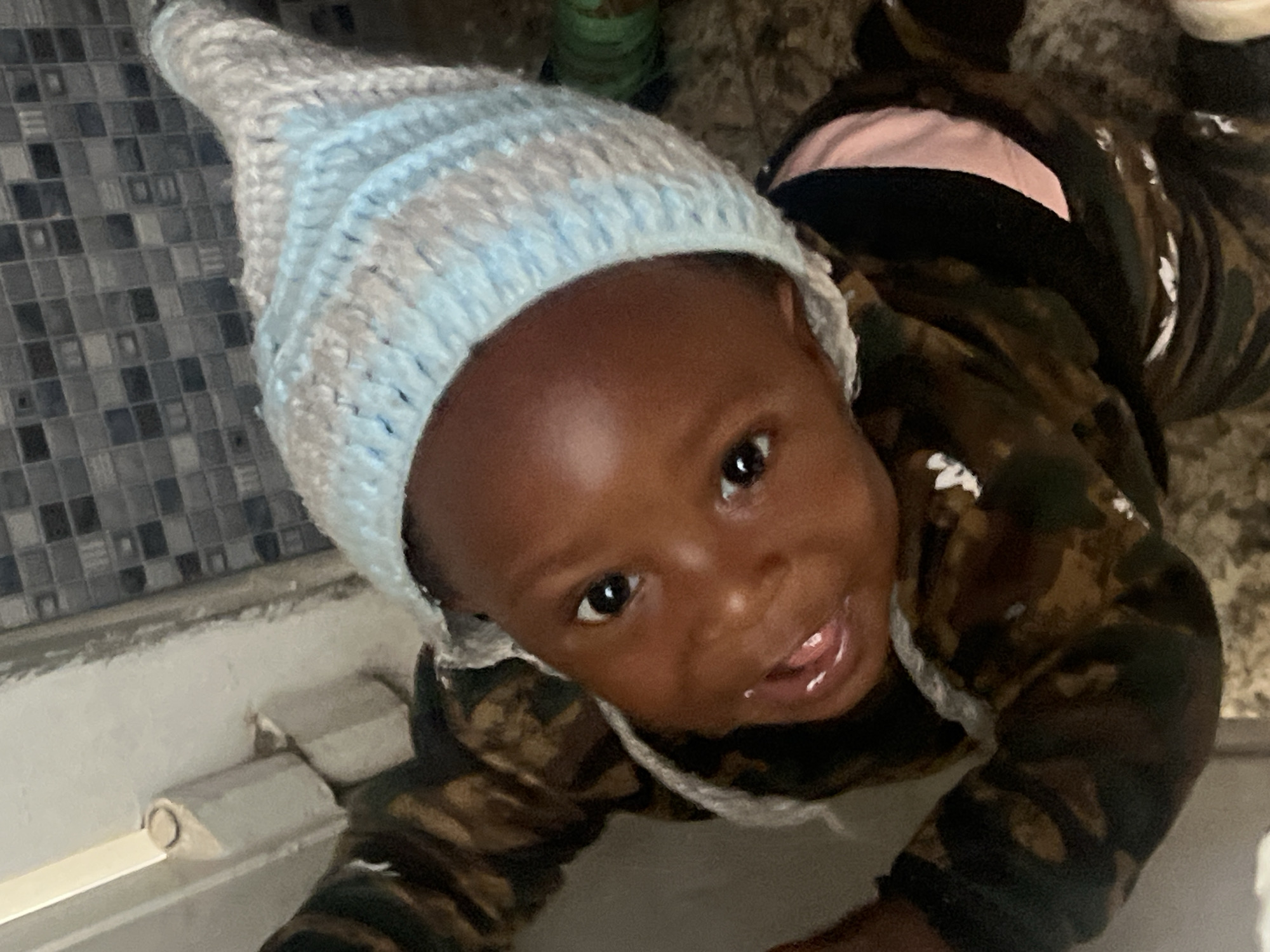 A baby is lying on a light-colored surface, possibly a floor, in an indoor setting. The baby is wearing a light blue and white knit cap that is slightly pointed. The baby is also wearing a long-sleeved shirt with a camouflage pattern. The floor in the background has a tiled pattern in muted colors. There is a white rectangular object near the lower left of the frame, possibly part of a baseboard or trim. The image is a close-up shot from a slightly high angle, looking down at the baby. The city of Gura Top, Nigeria is not discernible from the image.