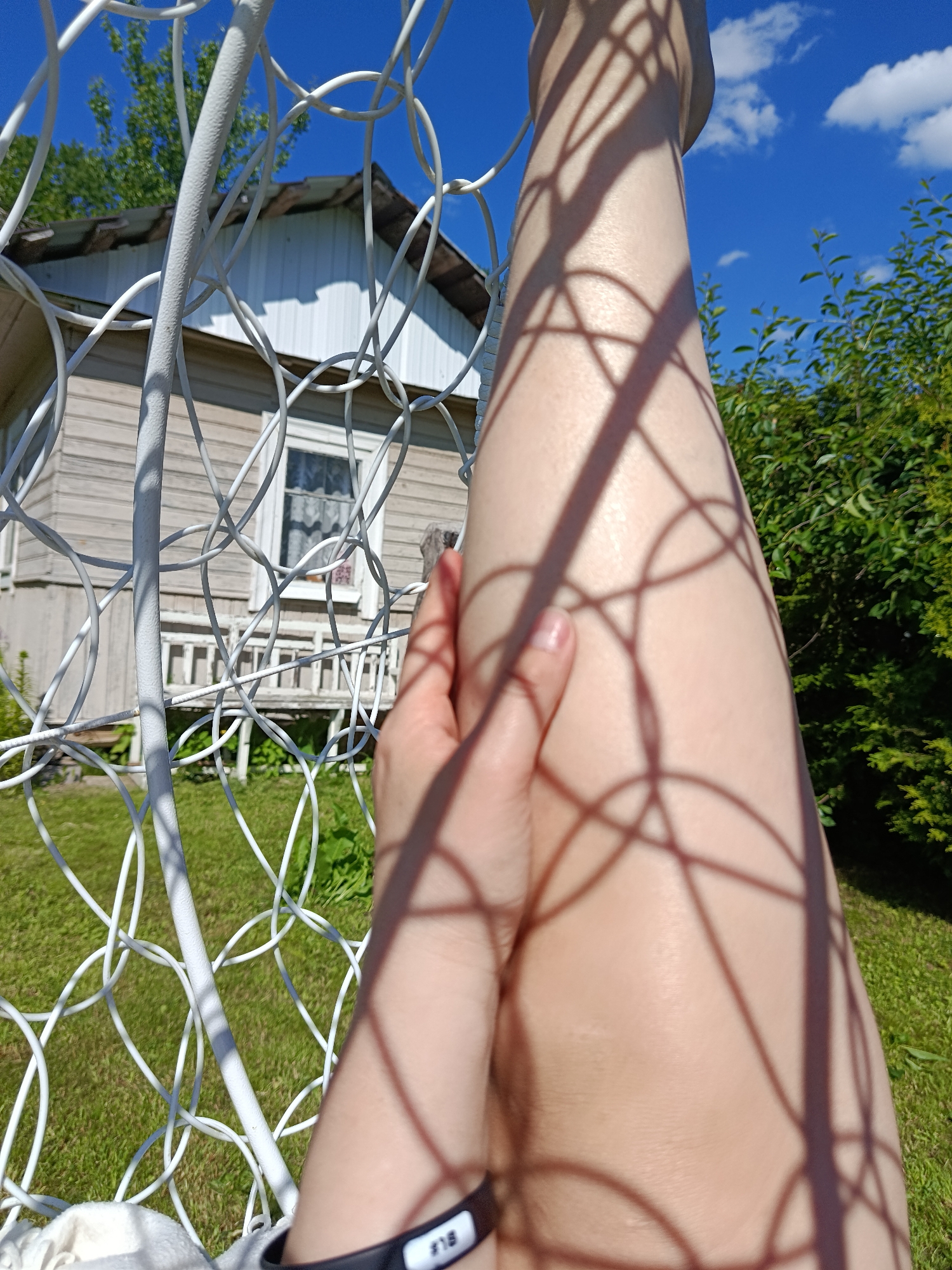 An overhead shot shows a person's bare legs and hand. The legs are tanned, and the hand is resting on the left leg. The scene is outdoors, with a blue sky and green trees. There's a white metal structure in the foreground, possibly a swing set. In the background, a wooden house with a white facade is visible. The location is the city of Shishkin Les, Russia.