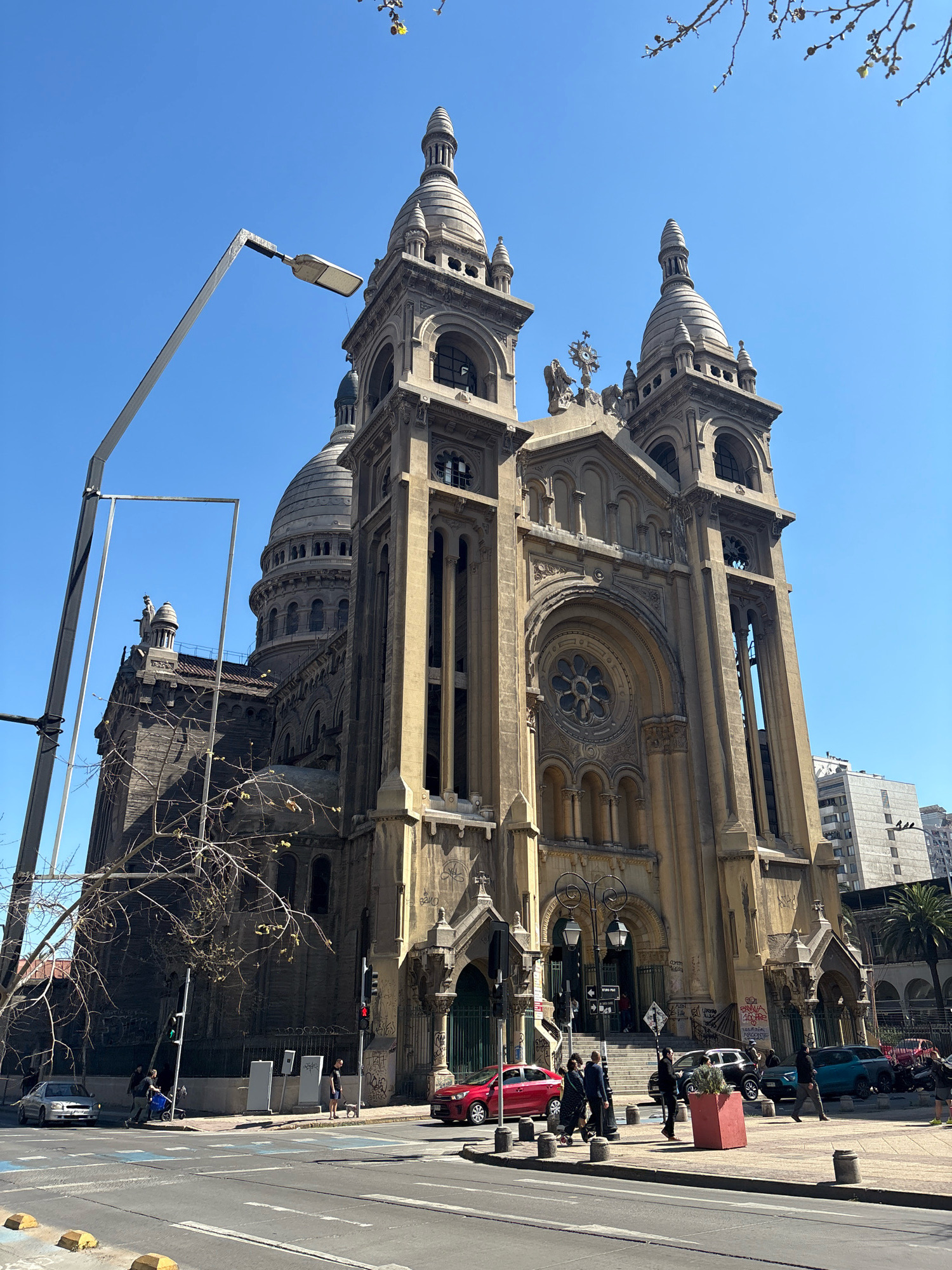 The image shows the imposing facade of the Basílica de la Gratitud Nacional in Santiago, Chile, bathed in sunlight under a clear blue sky. The Romanesque Revival basilica features multiple domes and spires, elaborate stonework, and arched windows. In the foreground, a street corner with pedestrian and vehicle traffic is visible. Several cars, including a red sedan and a silver hatchback, are on the street. Pedestrians walk along the sidewalk, some with shopping bags or pushing strollers. Traffic lights indicate the flow of traffic. A tall streetlamp with a modern, angular design stands prominently on the left side of the frame, partially obscuring the view of the basilica's left side. Bare tree branches in the middle ground suggest a temperate season. The background shows modern city buildings. The scene captures a typical urban streetscape in Santiago on a sunny day.